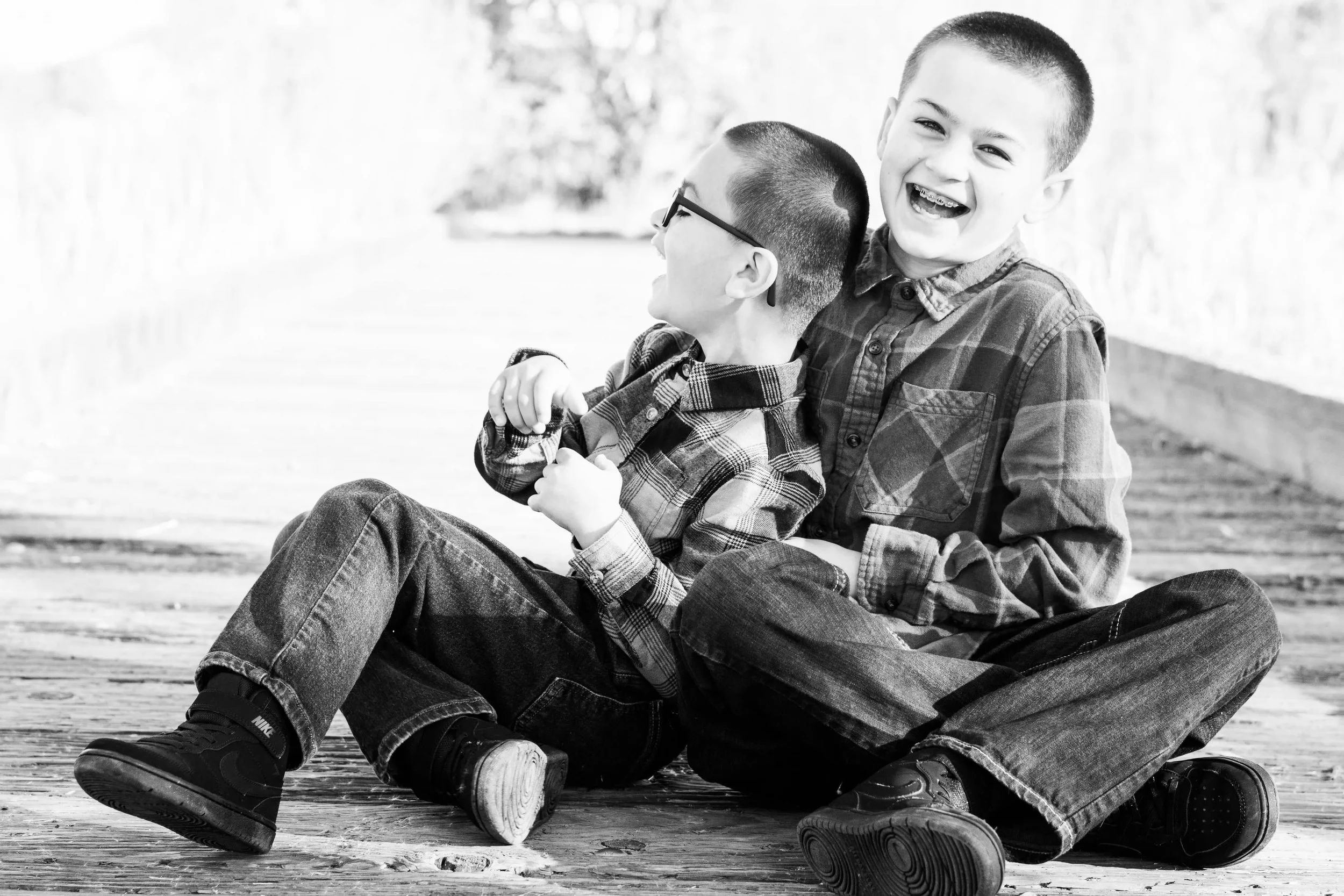 Two young boys sitting on a wooden surface outdoors, smiling and hugging each other, in black and white.