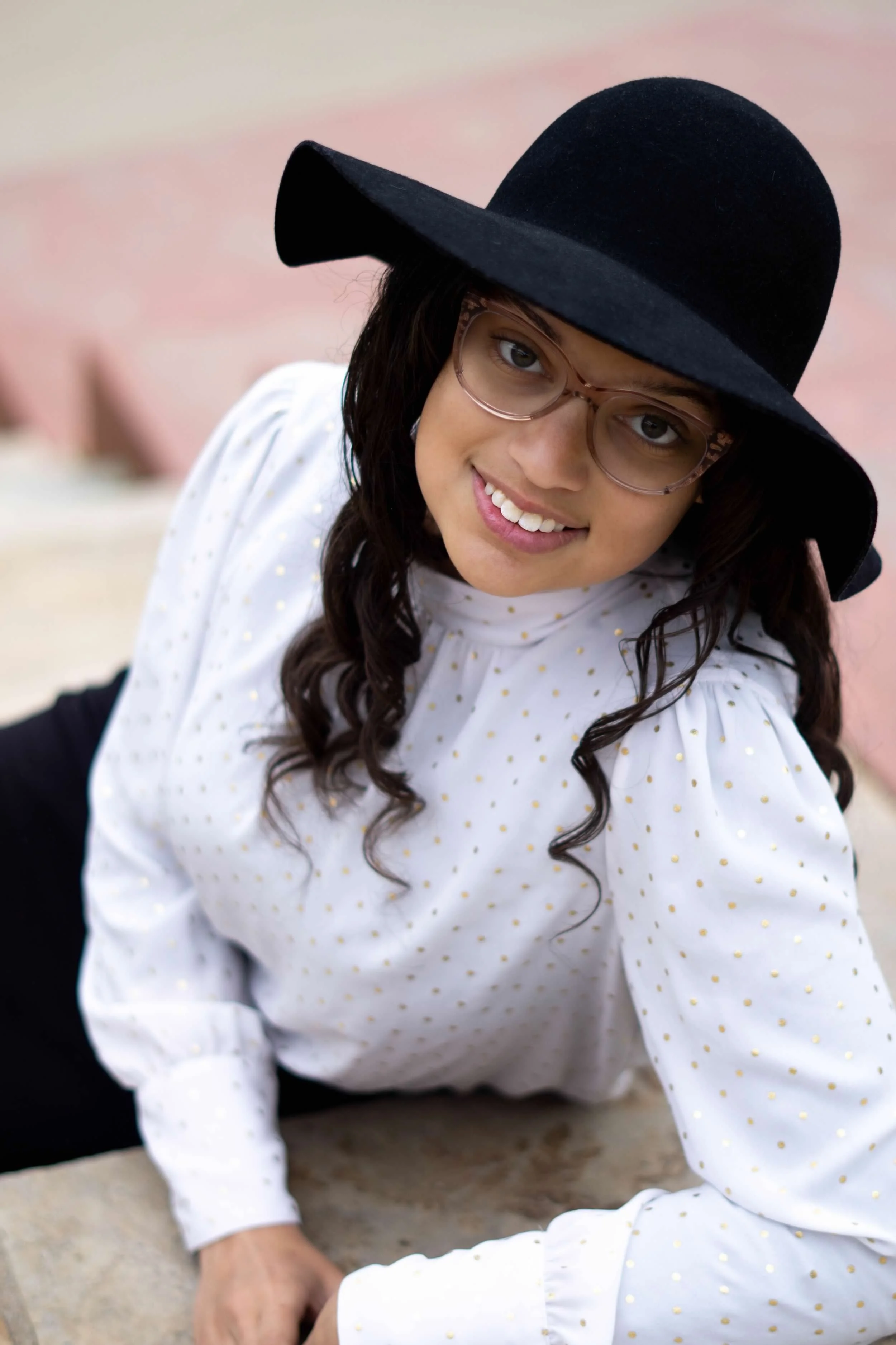 A woman smiling, wearing a black wide-brimmed hat, pink glasses, and a white dress with small yellow polka dots. She is seated on the ground outdoors.