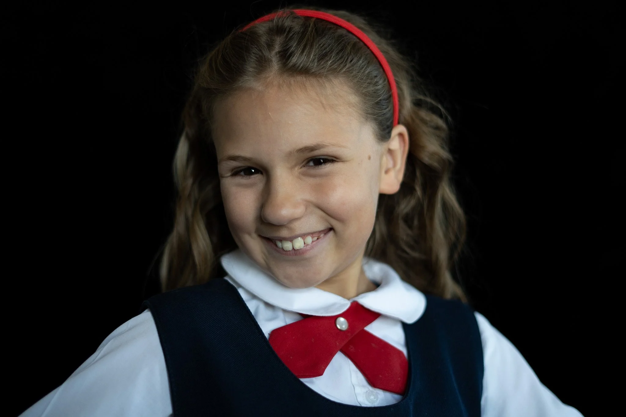 Young girl with light brown hair, wearing a red headband, school uniform, and a backpack, smiling against a black background.