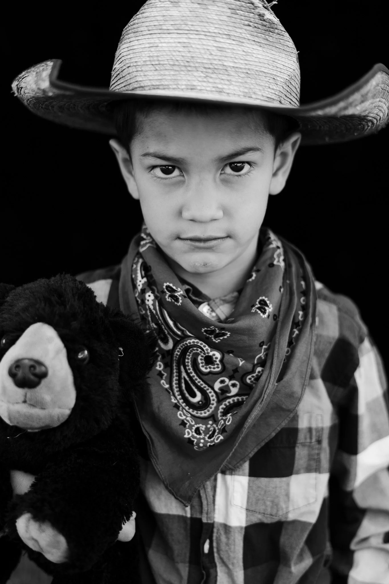 A young boy wearing a cowboy hat and bandana, holding a plush toy, dressed in a plaid shirt with a background of other children or people.