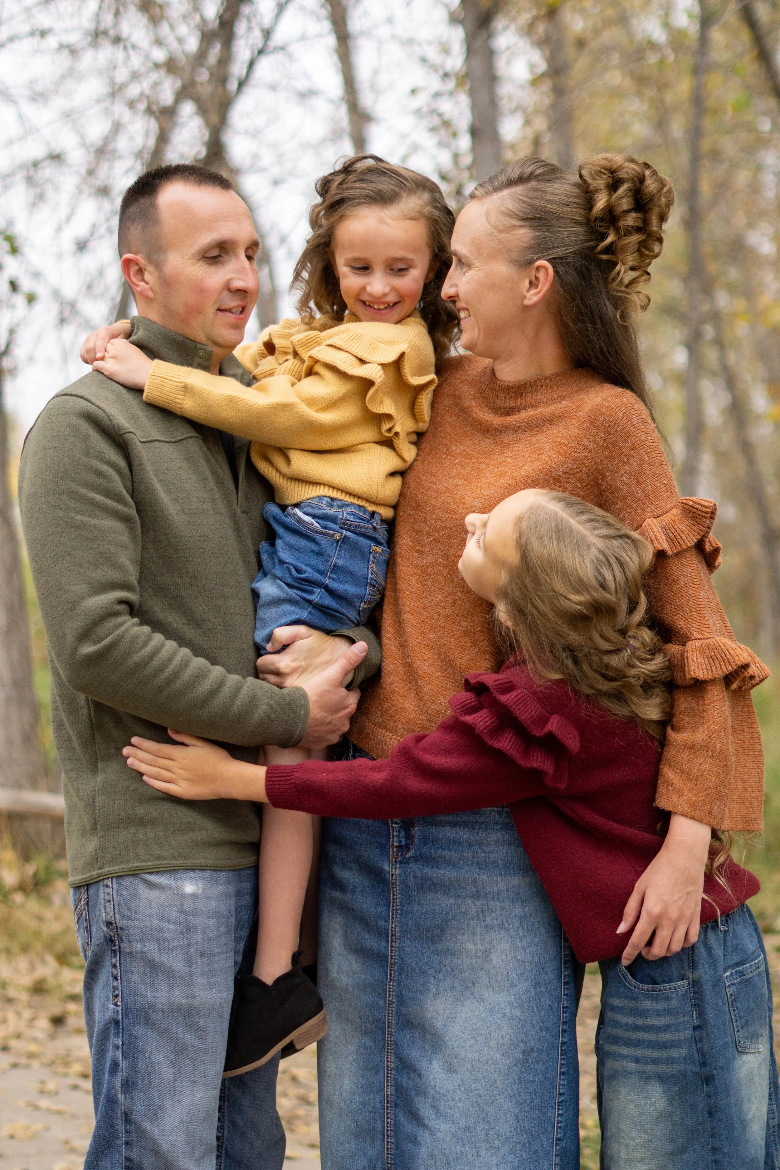 A family of four, including a man, a woman, and two young girls, smiling and hugging outdoors in a park with autumn trees.