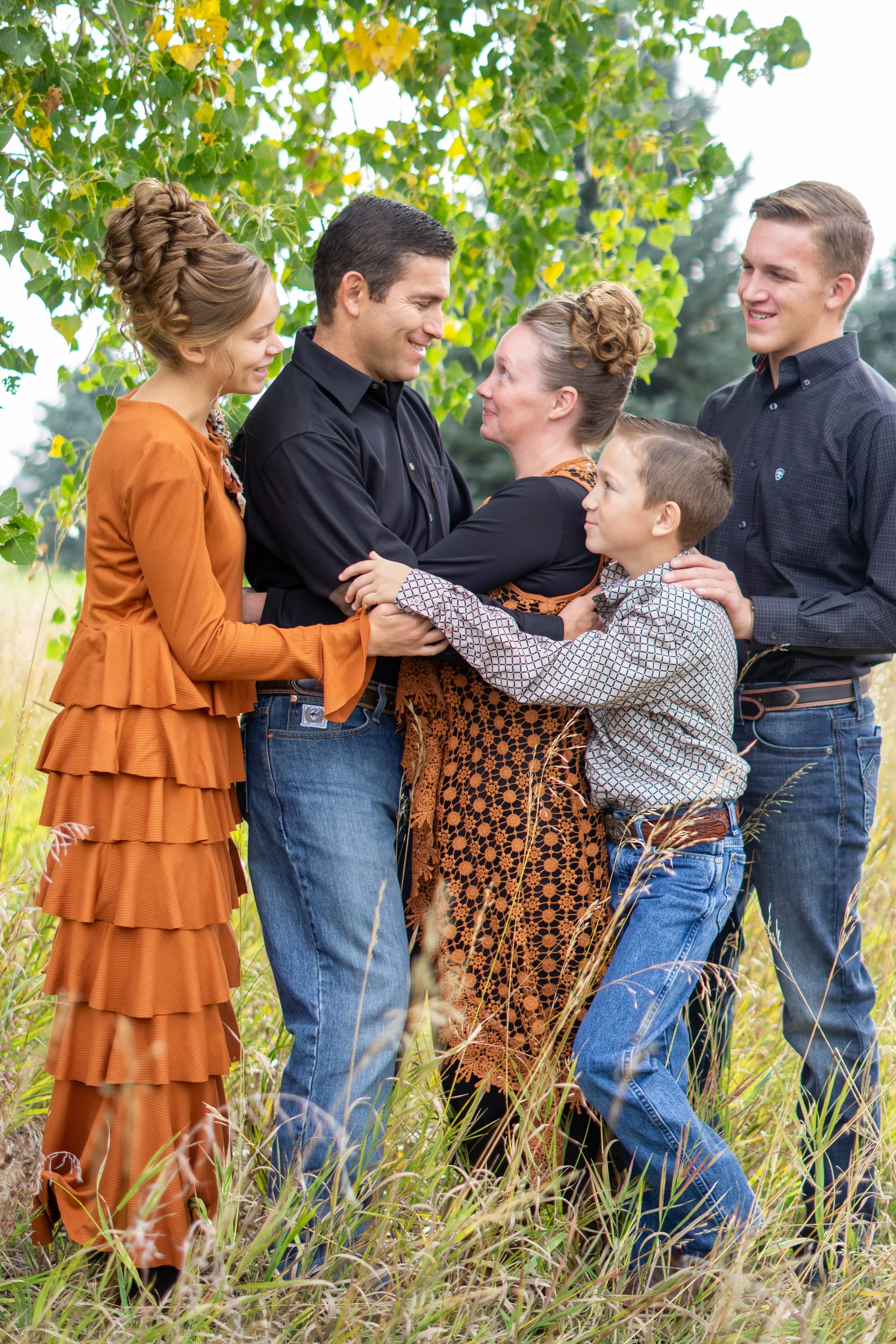 Family of six holding hands and smiling together outdoors in a grassy field with green trees in the background, celebrating a happy moment.