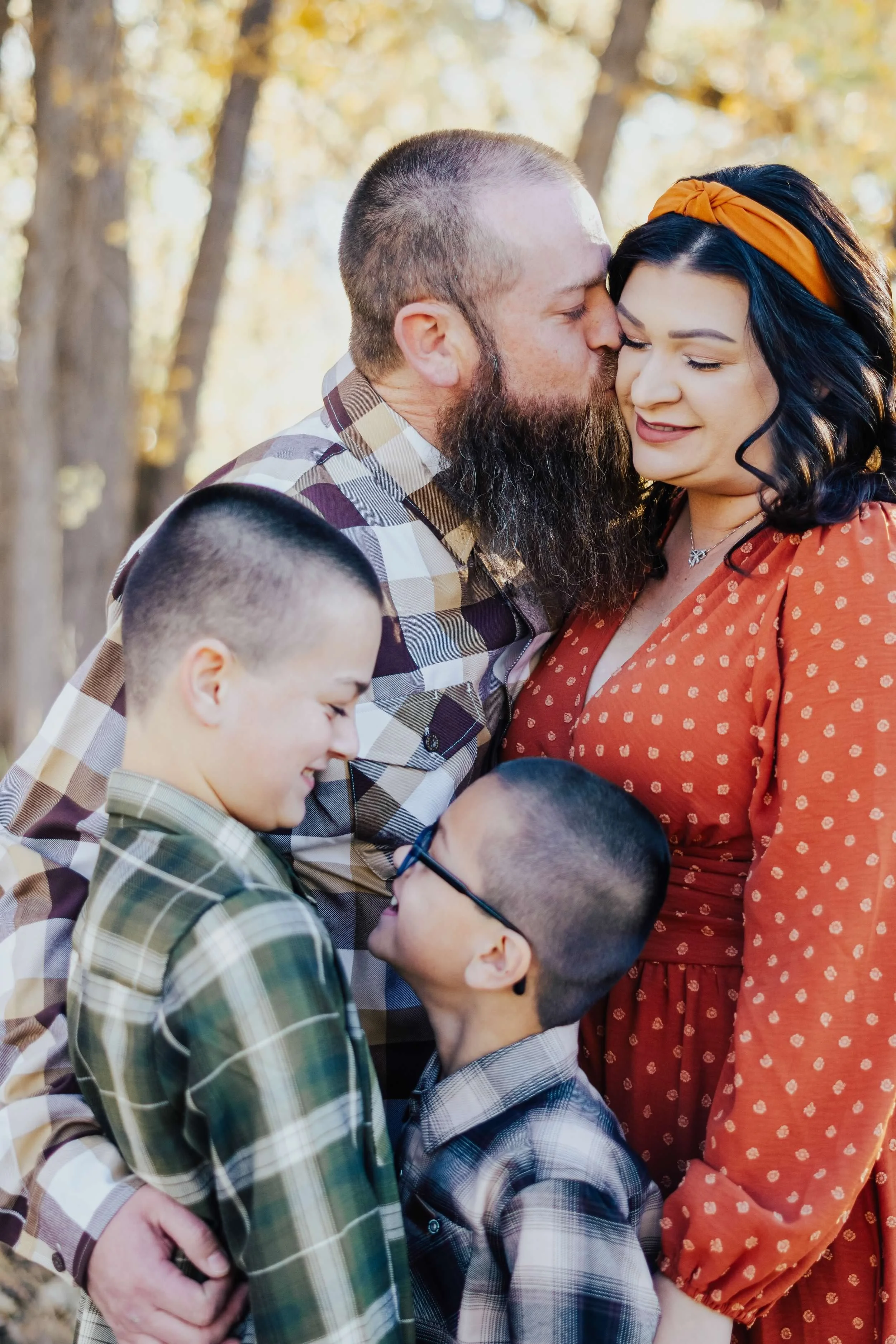 Family gathering outdoors during autumn, with three children and two adults sharing a close moment with kisses and smiles, surrounded by fall foliage.