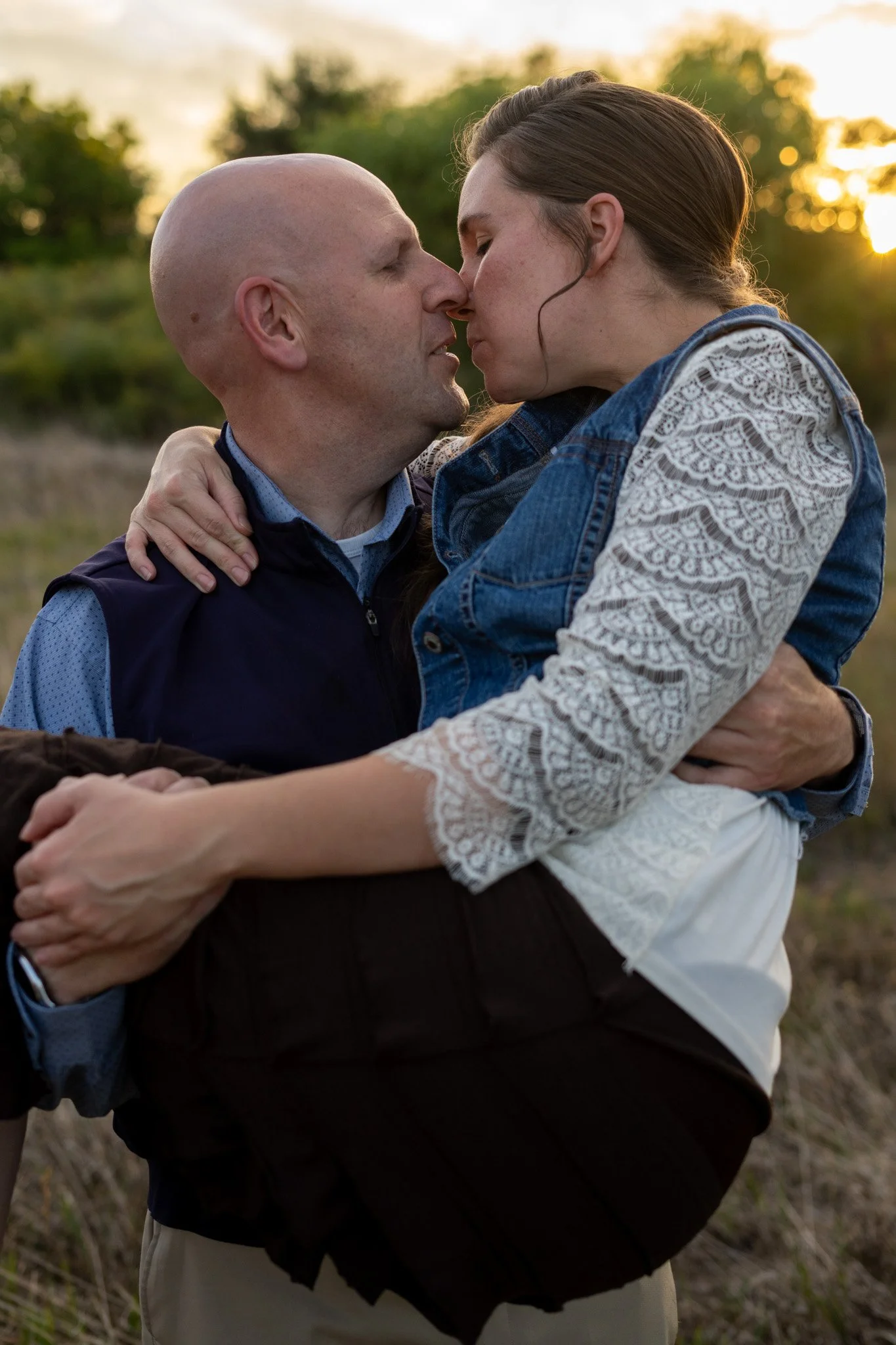 Two people, a man and a woman, are hugging outdoors during sunset. Their faces are very close, and the man has his eyes closed while the woman is gently holding him, both showing affection. The background features trees and a warm, golden sky.