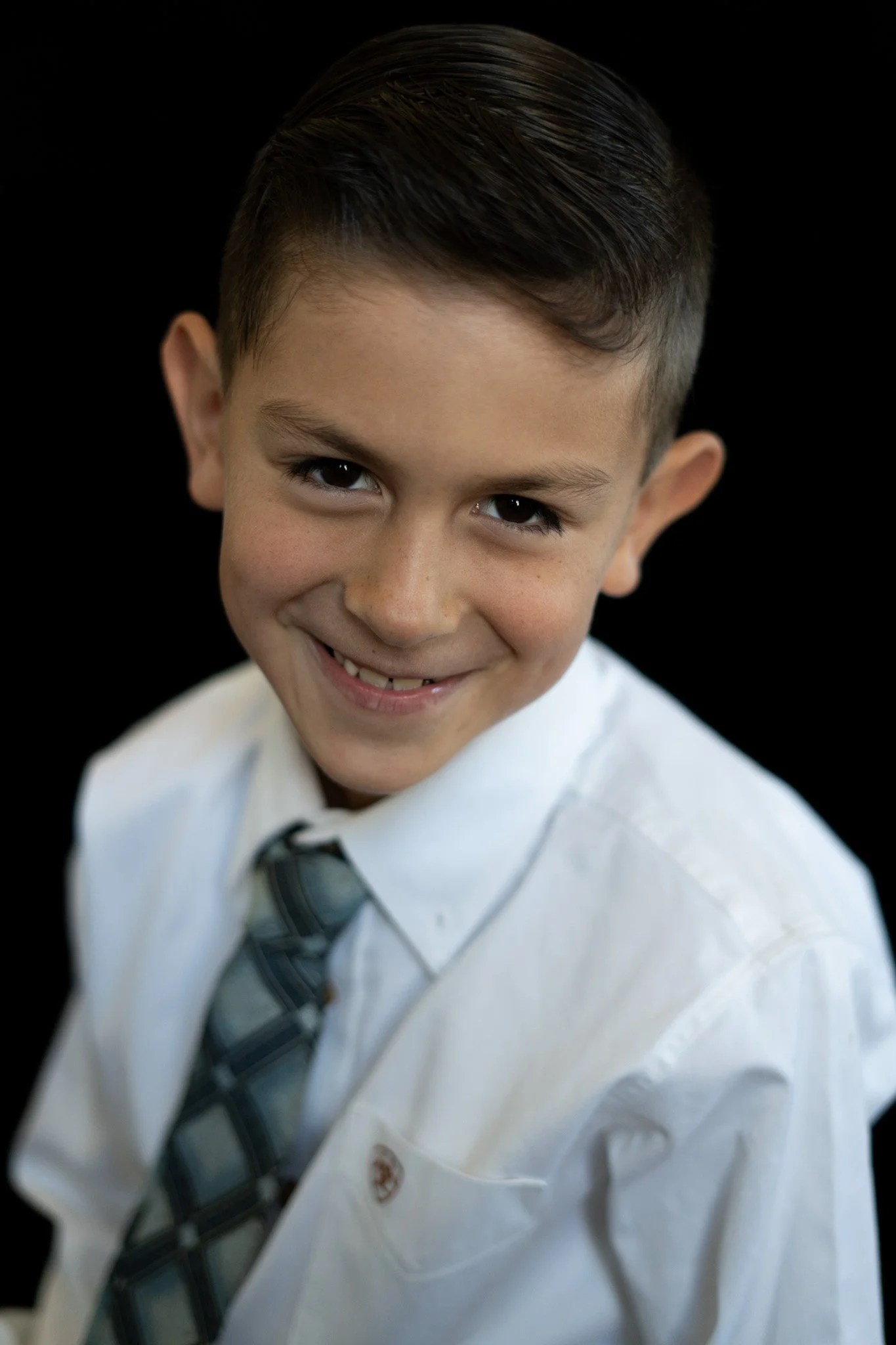 Close-up of a young boy in a white shirt and tie, smiling with dark hair styled neatly, against a black background.