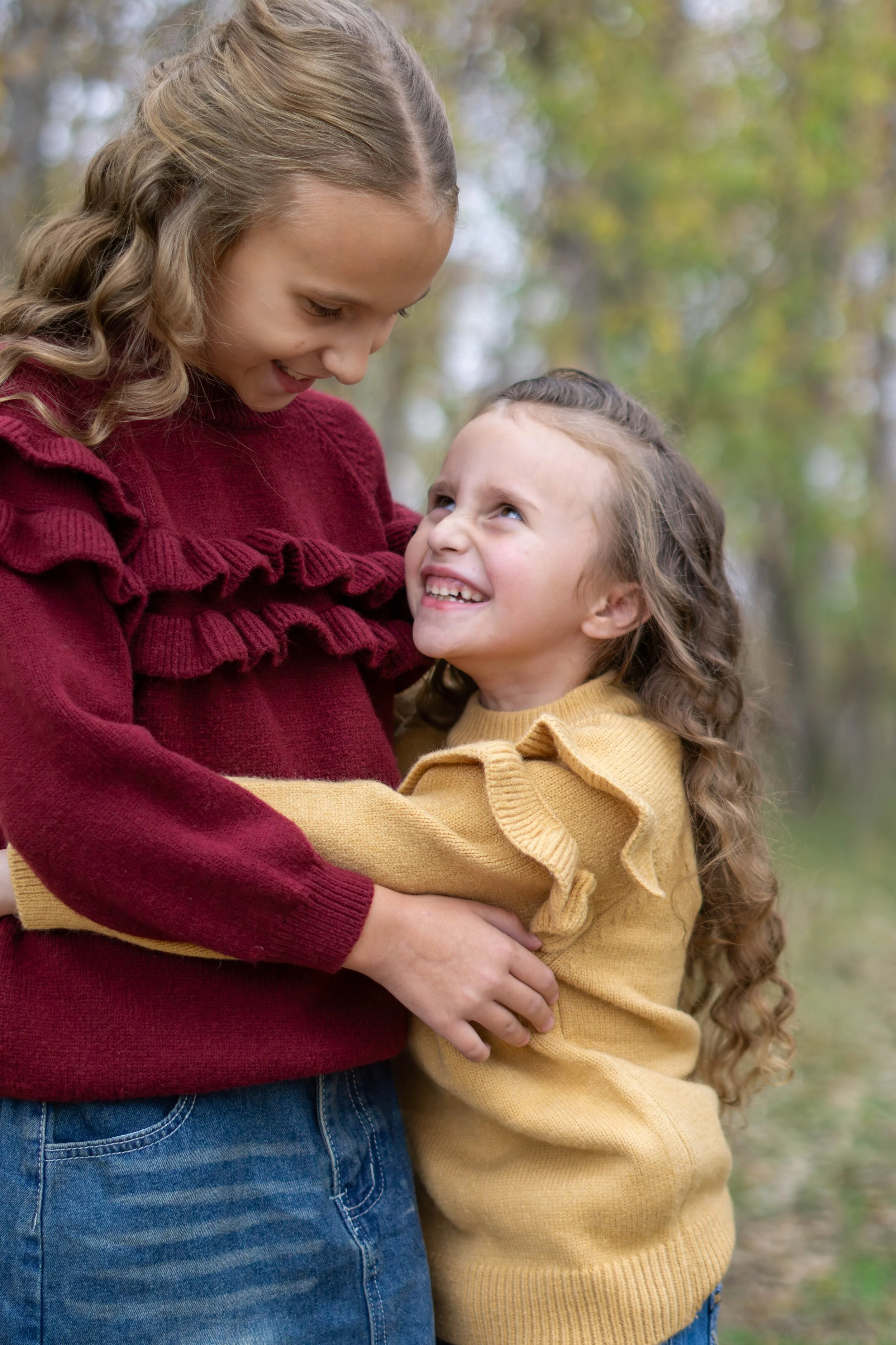 A woman and a young girl are outdoors, hugging each other and looking at each other affectionately. The woman is wearing a burgundy sweater and jeans, while the girl is wearing a mustard yellow sweater. The background features trees with autumn foliage.