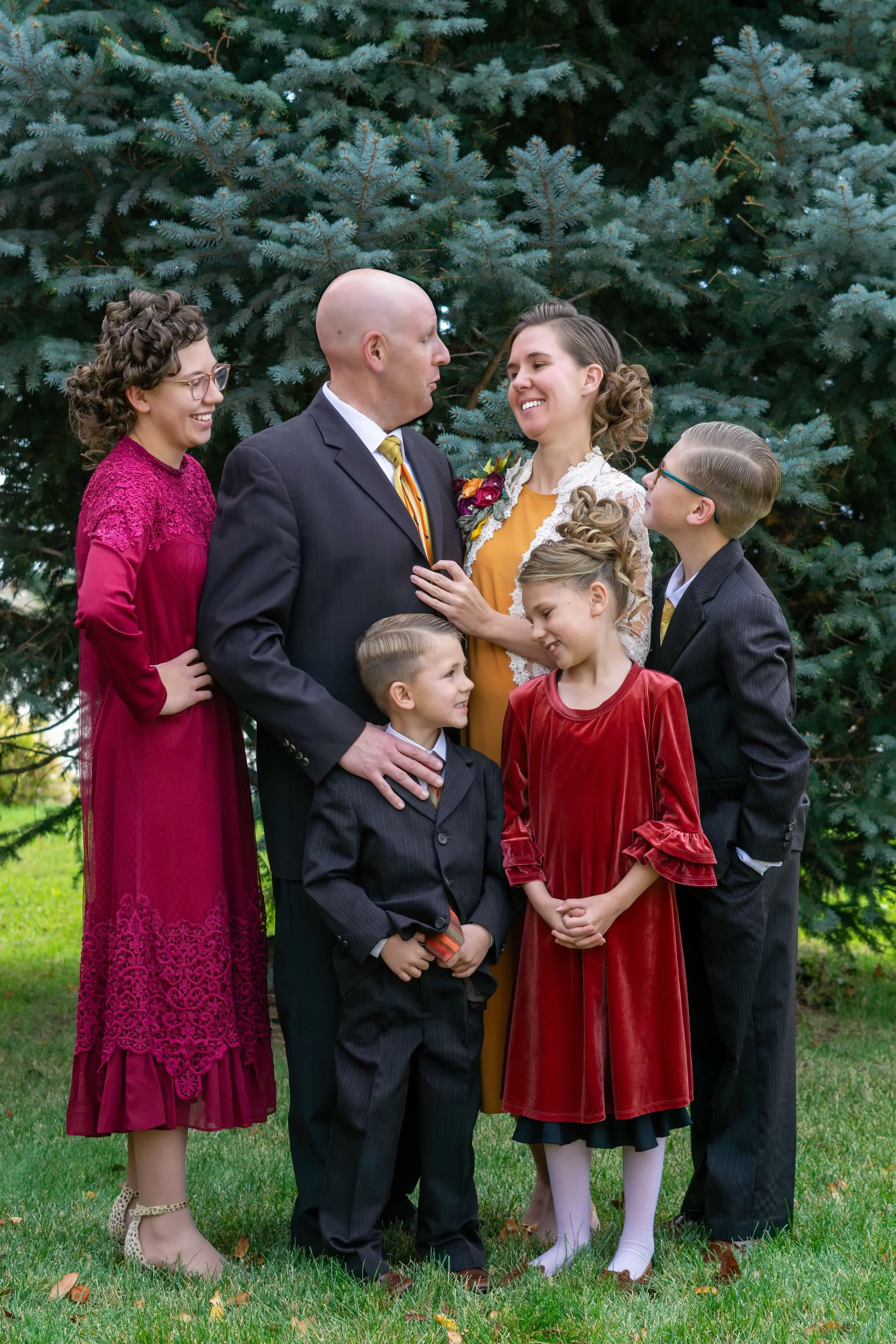A family of six standing on grass, dressed in formal and semi-formal attire, in front of pine trees. The group includes two adult men and women, and four children, all smiling and engaging with each other.