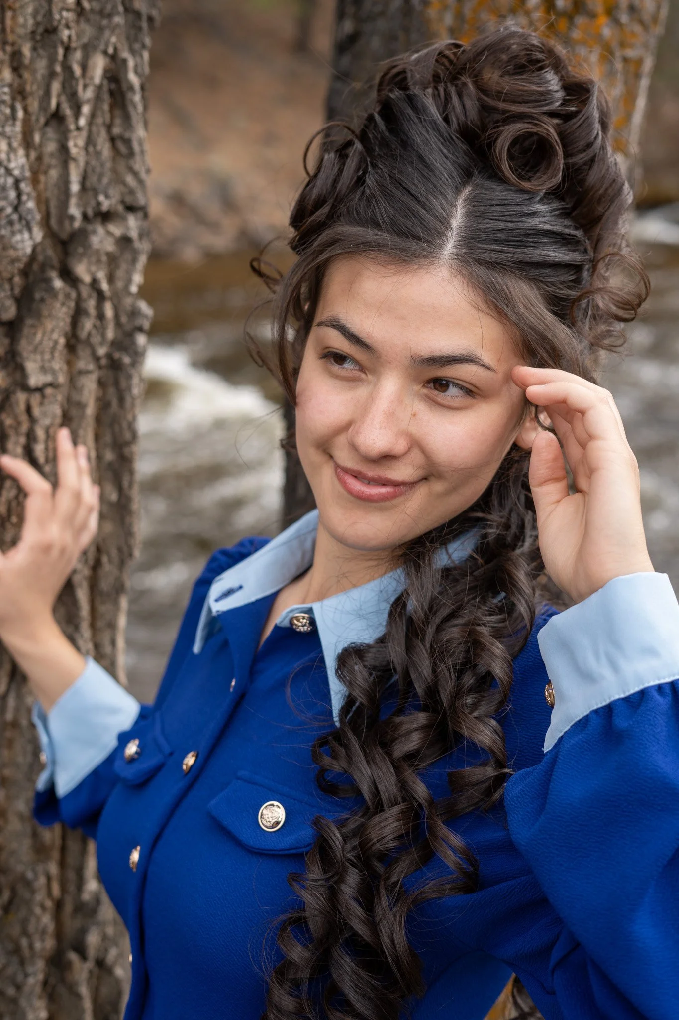 A woman with long curly dark hair styled in an updo, wearing a blue jacket with gold buttons, standing outdoors near a tree and water in the background.