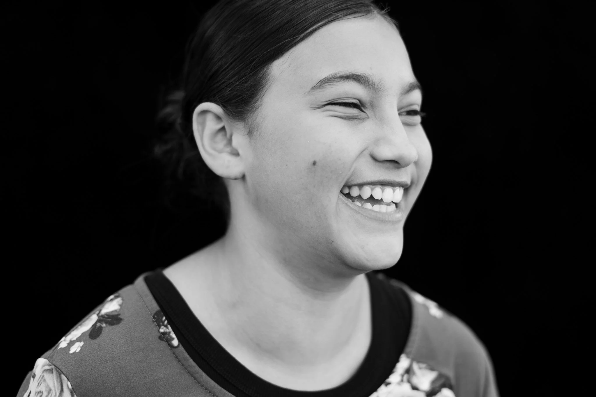A young woman with dark hair smiling and winking, wearing a floral top against a black background.