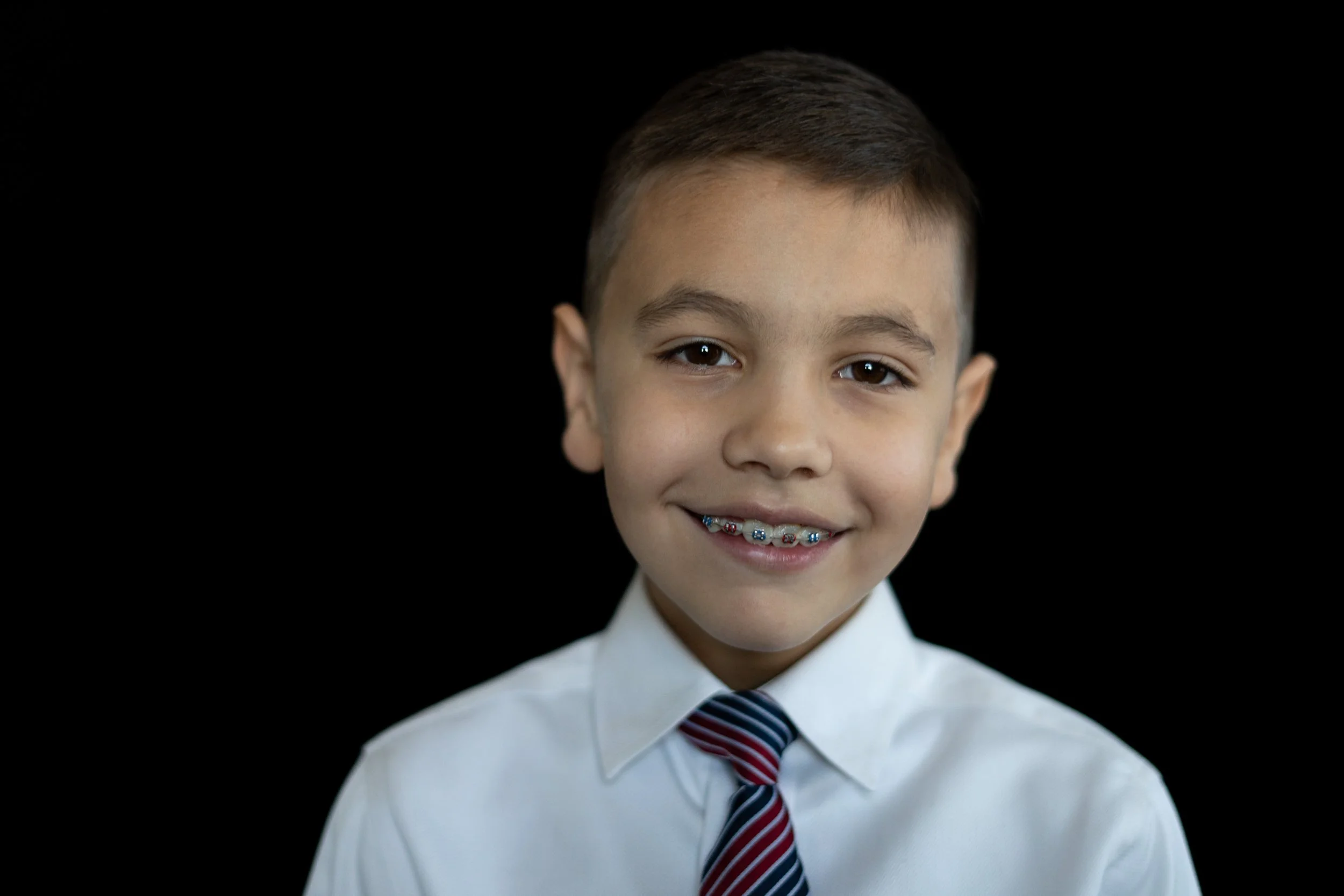 A young boy with short brown hair, wearing a white shirt and a striped tie, smiling with braces on his teeth, against a black background.
