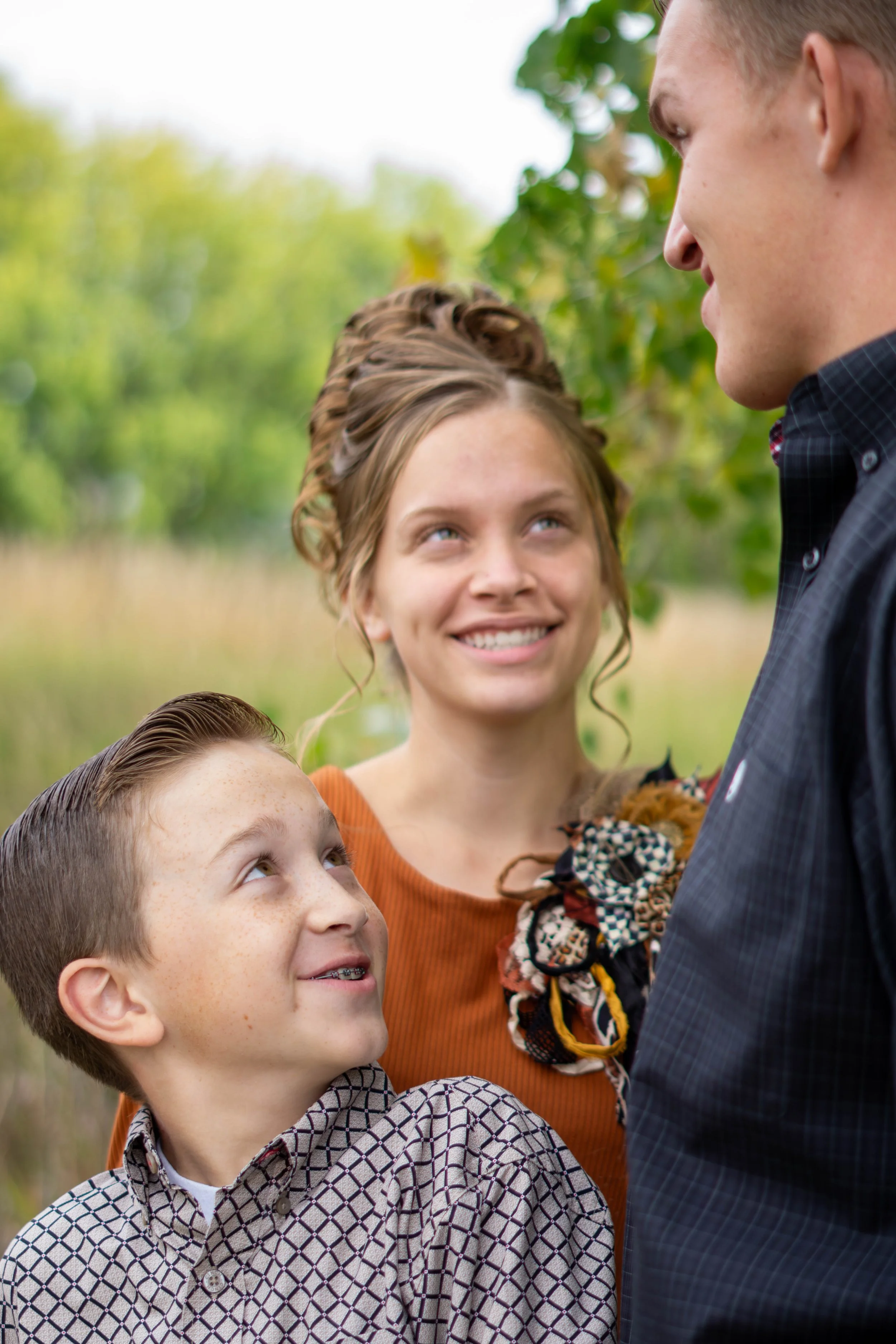 A group of people outdoors, including a woman with curly hair smiling and looking up at a man, a young boy looking up at the man, and a girl with braided hair, all facing a man dressed in a dark suit.