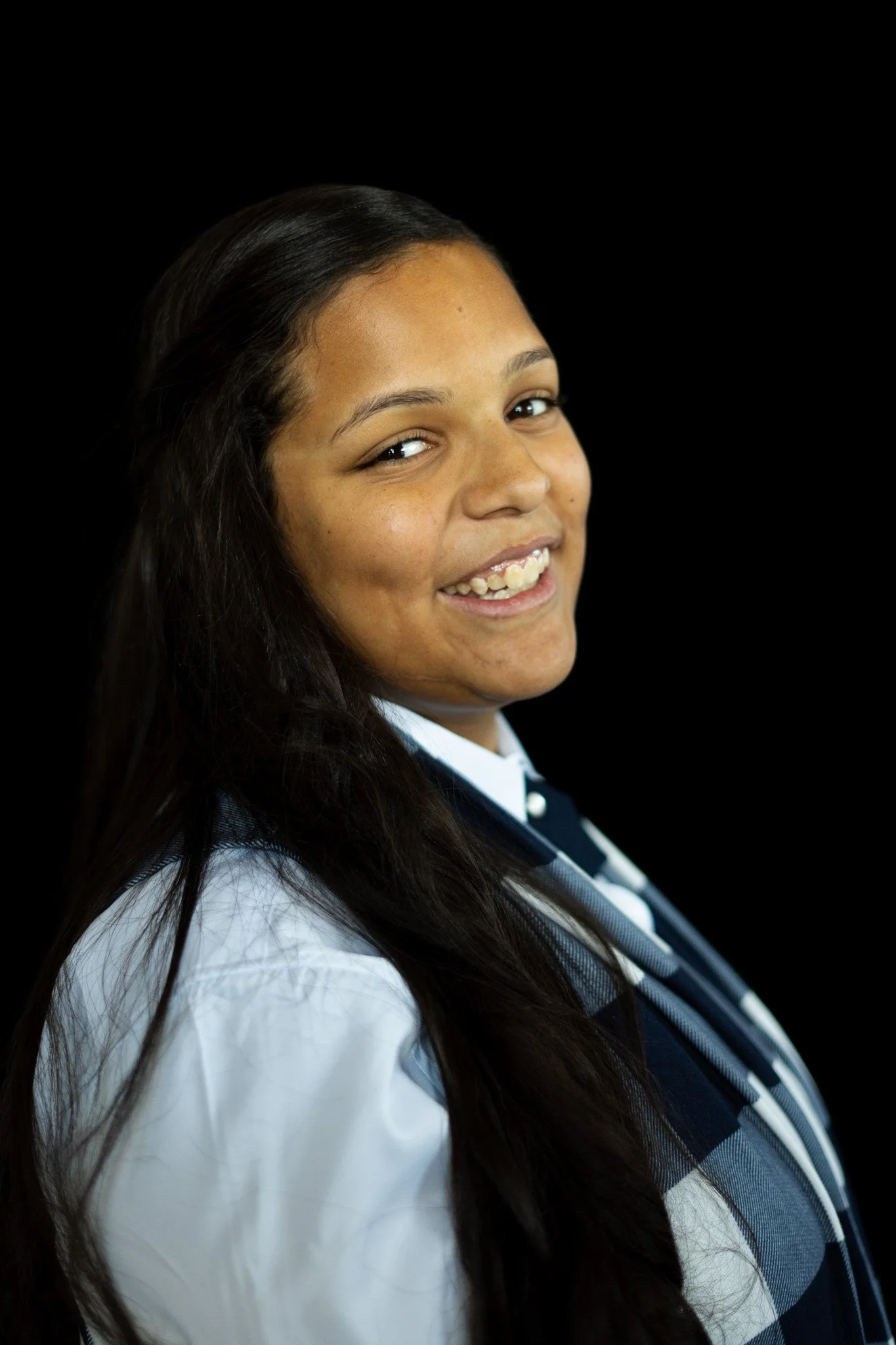 Young woman with long dark hair smiling, wearing a school uniform with a navy and white blazer and a white shirt, against a black background.