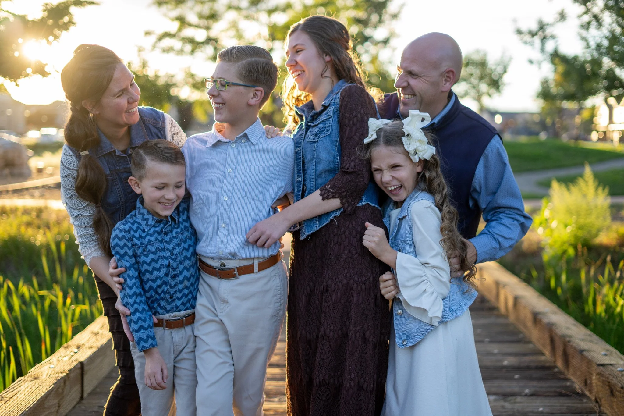 A family of six, including three children and three adults, standing on a wooden footbridge outdoors during sunset, smiling and laughing together.