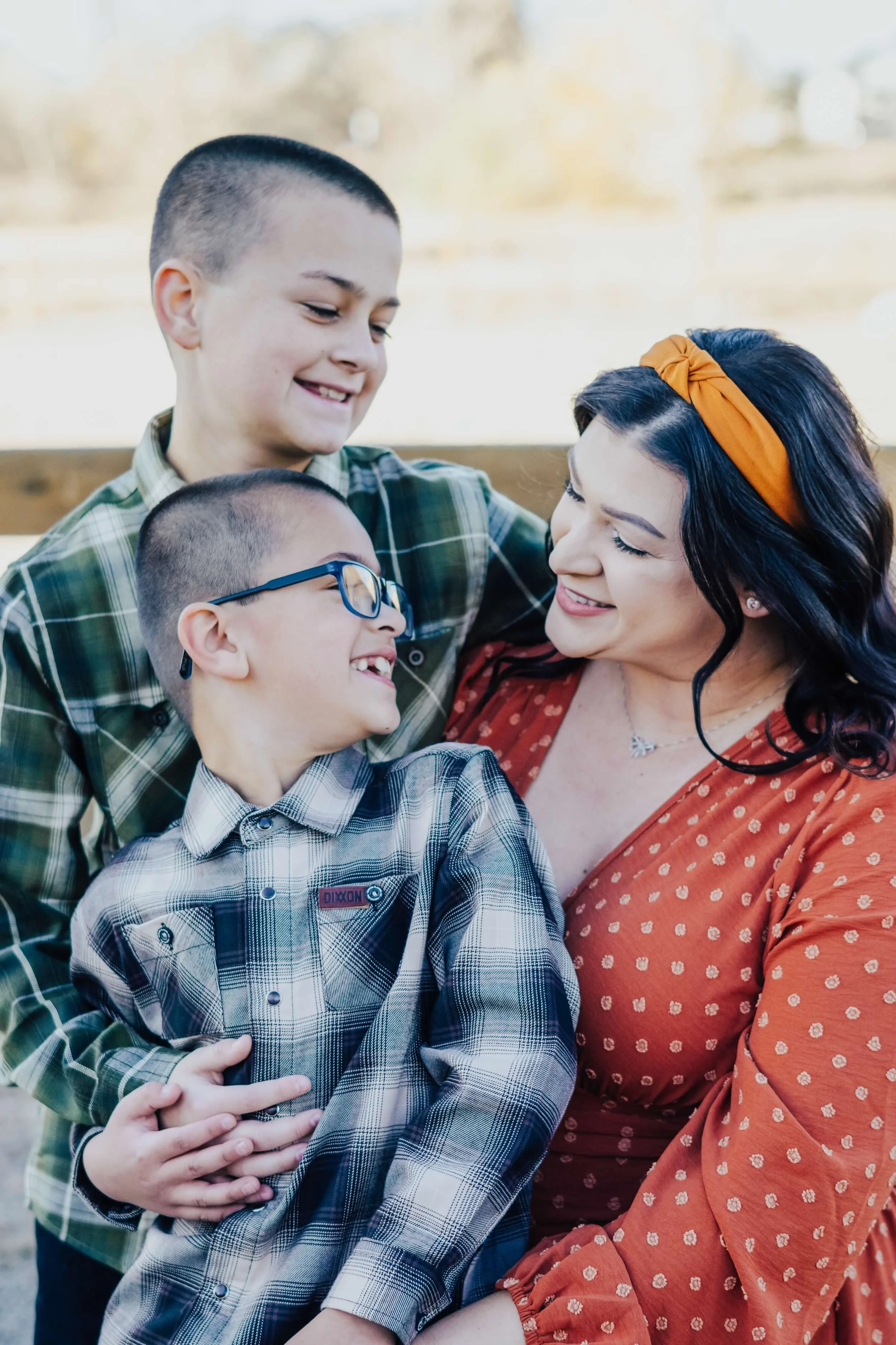 A woman with dark hair, wearing an orange headband and a red dress with white polka dots, smiling and looking at two young boys, one with glasses and the other wearing a green plaid shirt, as they all enjoy a moment together outdoors.
