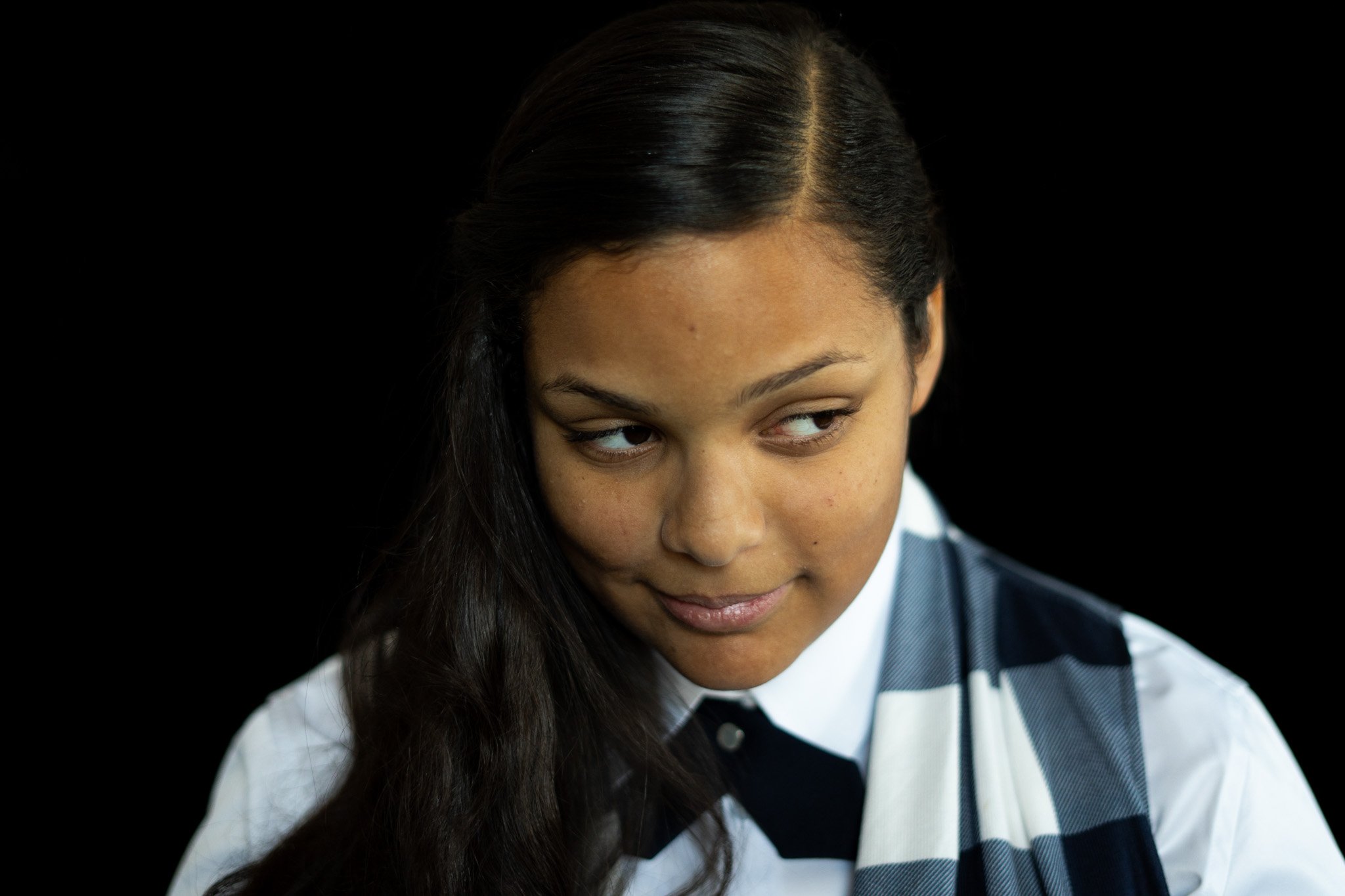 A young woman with long dark hair, wearing a shirt, tie, and vest, looking to her left with a slight smile against a black background.