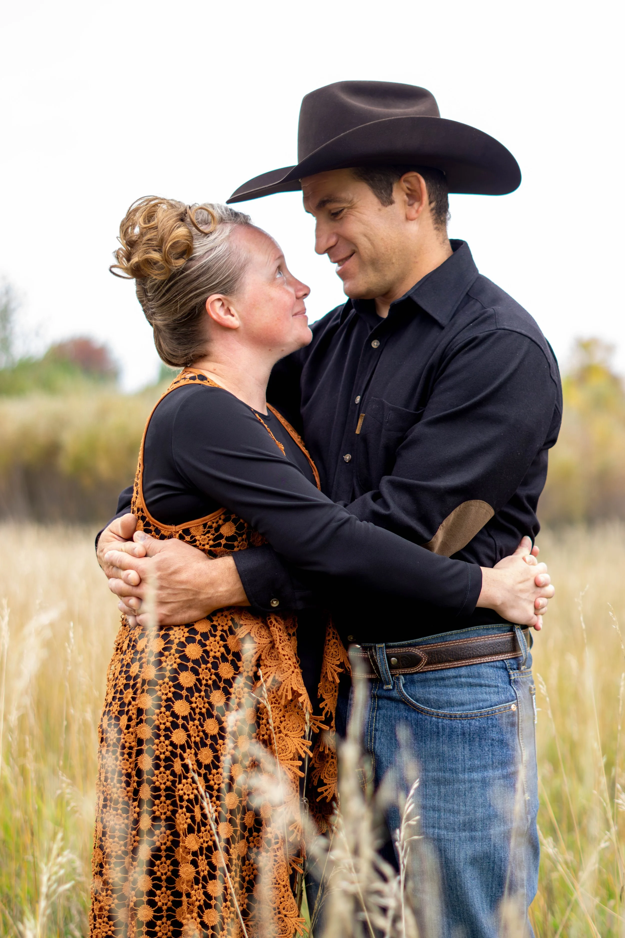 A man and woman are embracing each other in a field, smiling and looking into each other's eyes. The woman has curly blonde hair styled in an updo, and the man is wearing a black cowboy hat and a dark button-up shirt.