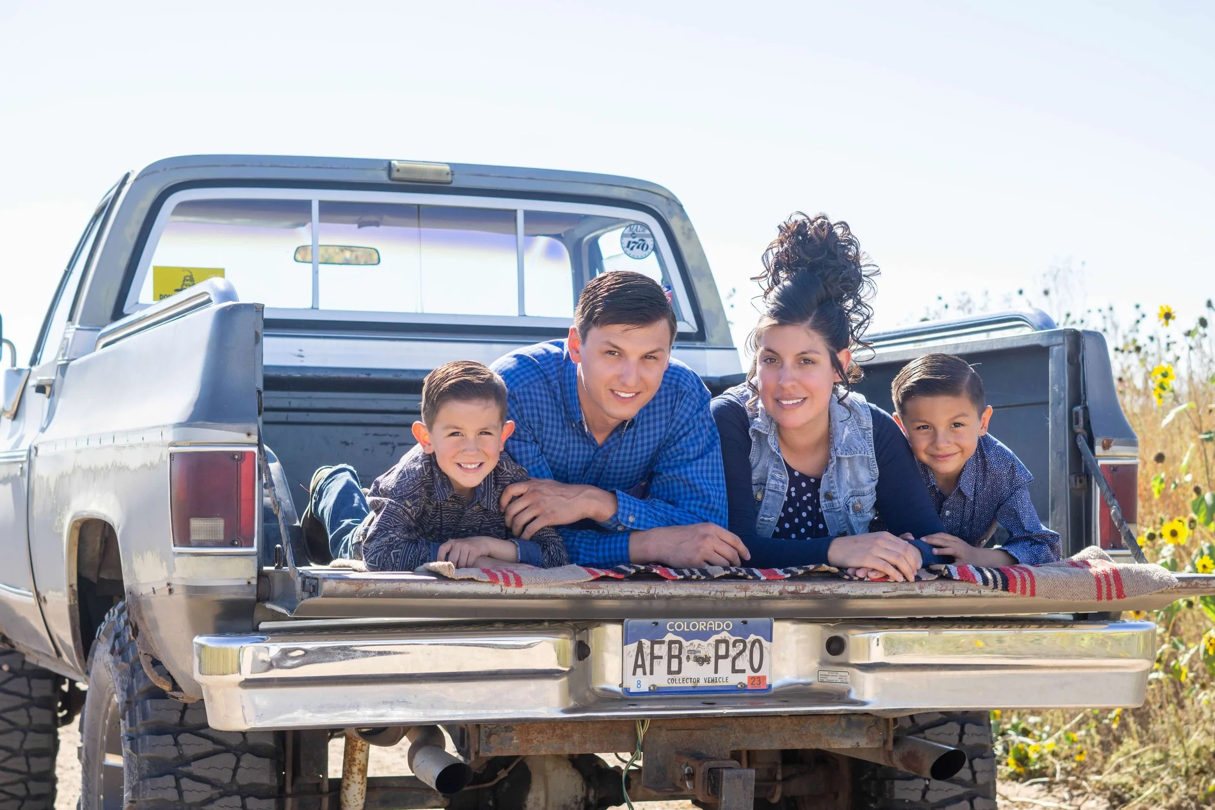 A family of five sitting in the open tailgate of a pickup truck in a countryside setting, with a field of sunflowers in the background. They are smiling at the camera, with two children and two adults.