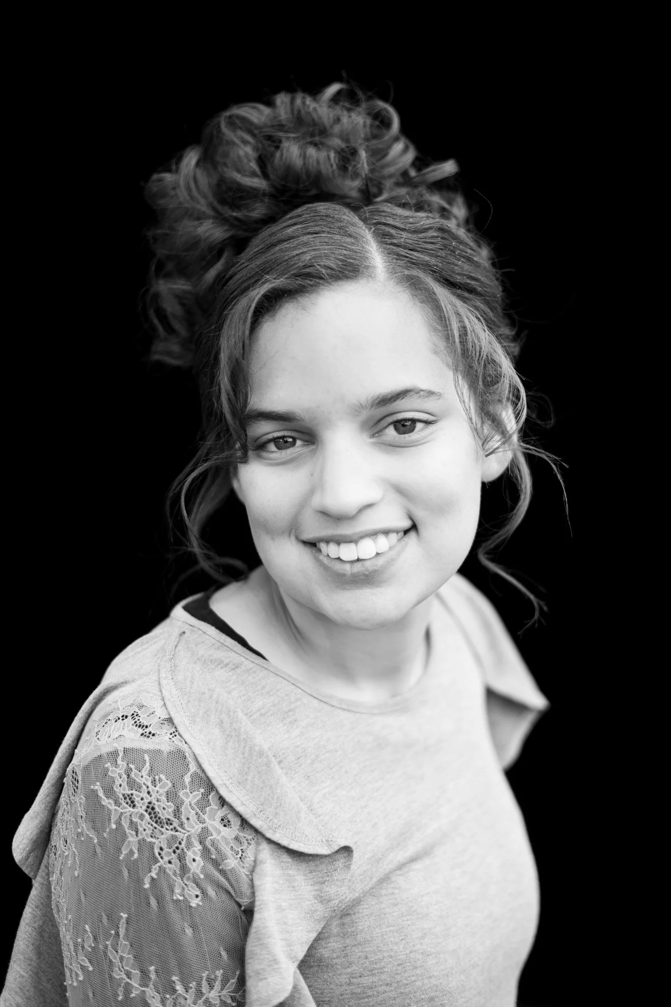 Black and white photo of a smiling young woman with curly hair styled in a bun, wearing a lace-sleeved top.