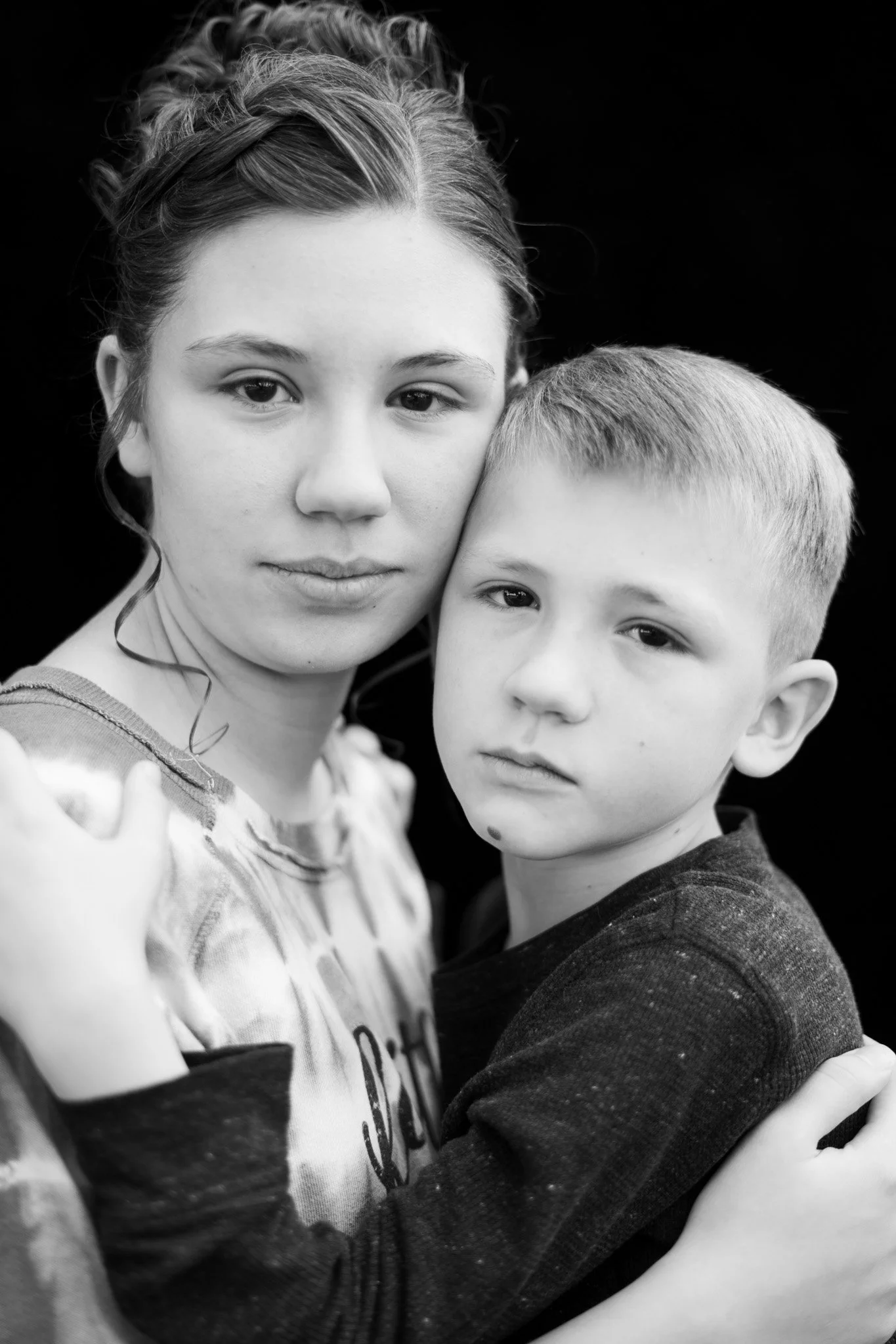A black and white photo of a woman and a young boy embracing each other. The woman has her eyes slightly open and is looking directly at the camera, with her hair pulled back. The boy has short hair and a serious expression, looking at the camera as well. They are holding each other closely against a plain, dark background.