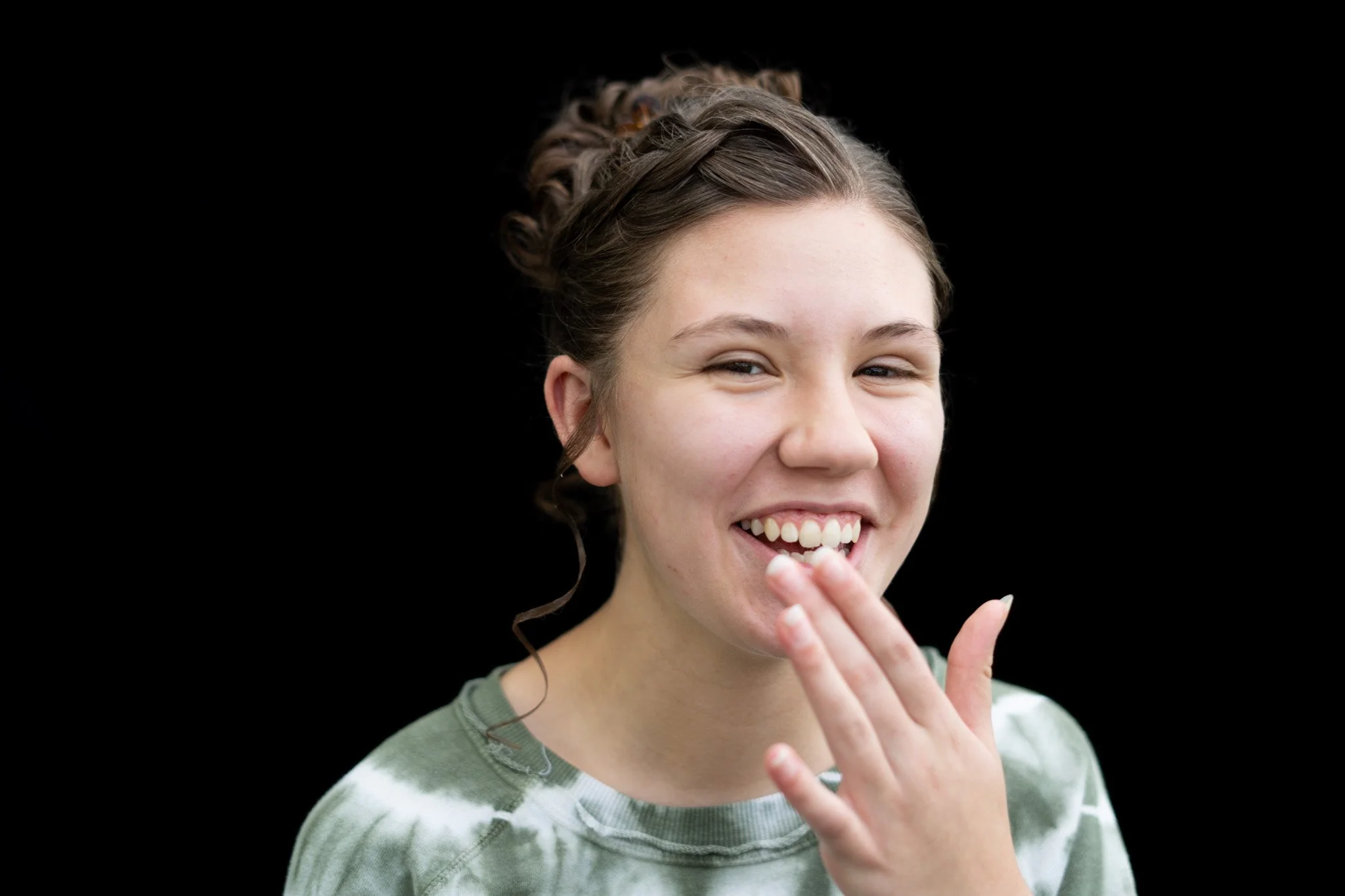 A young woman with light skin and dark hair styled in an updo, smiling and covering her mouth with her hand, wearing a green and white striped shirt.