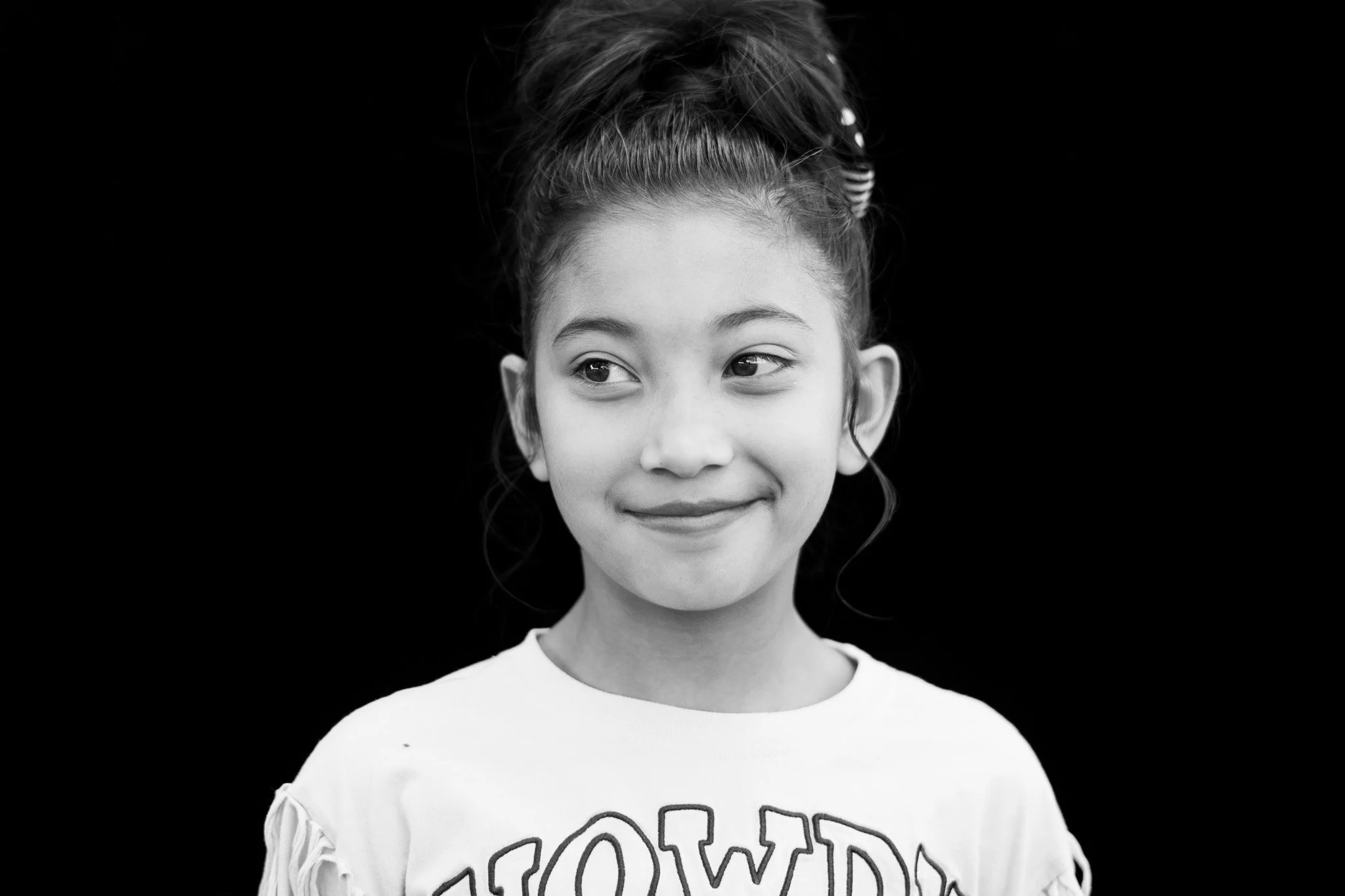 Black and white photo of a young girl with a happy expression, wearing a shirt with partially visible text, against a black background.