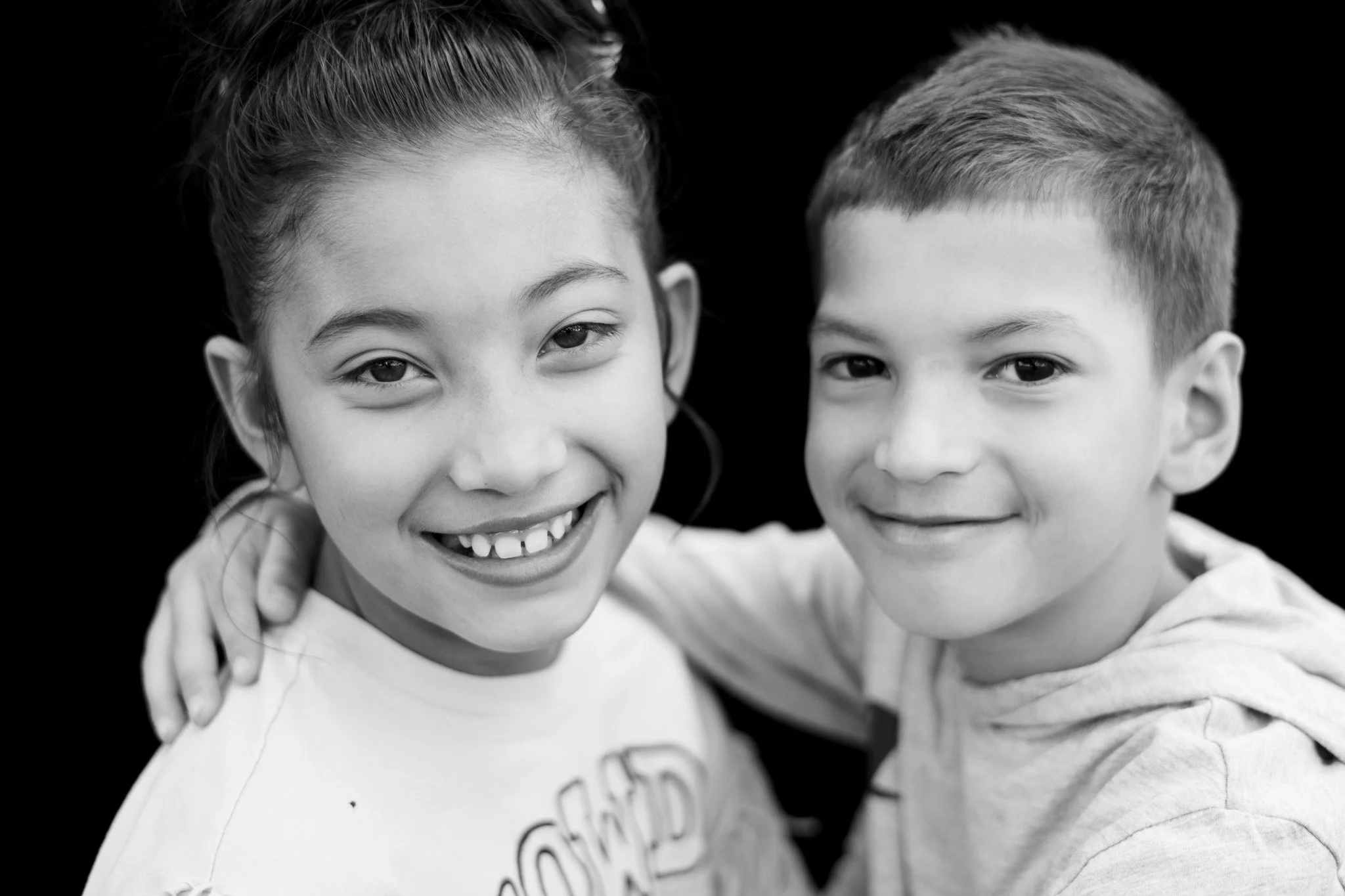 A black and white photo of a young girl and boy smiling with their arms around each other, against a dark background.