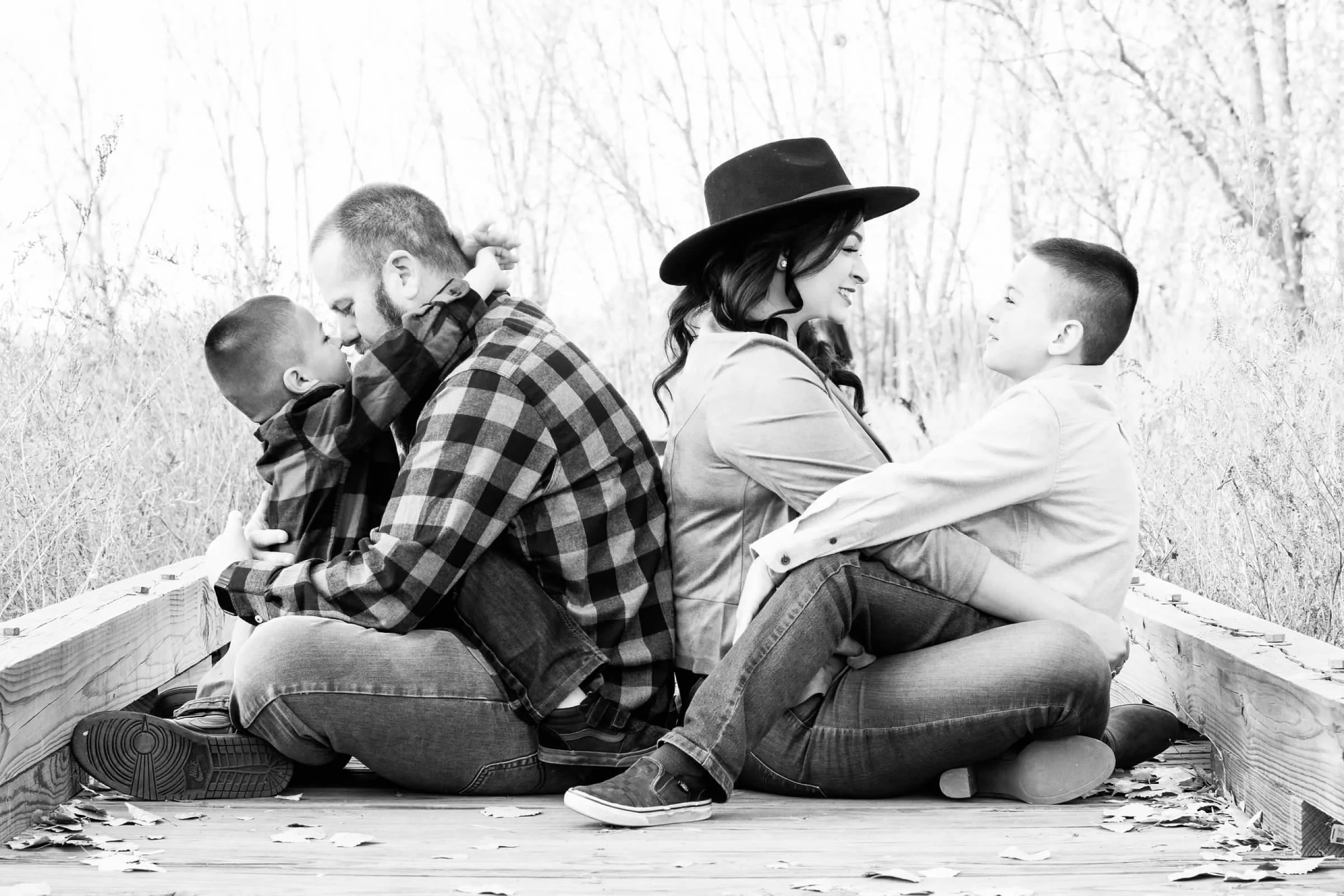 A black and white photo of two families sitting on a wooden bridge outdoors, facing each other and smiling, with two children and adults in a natural setting with trees.