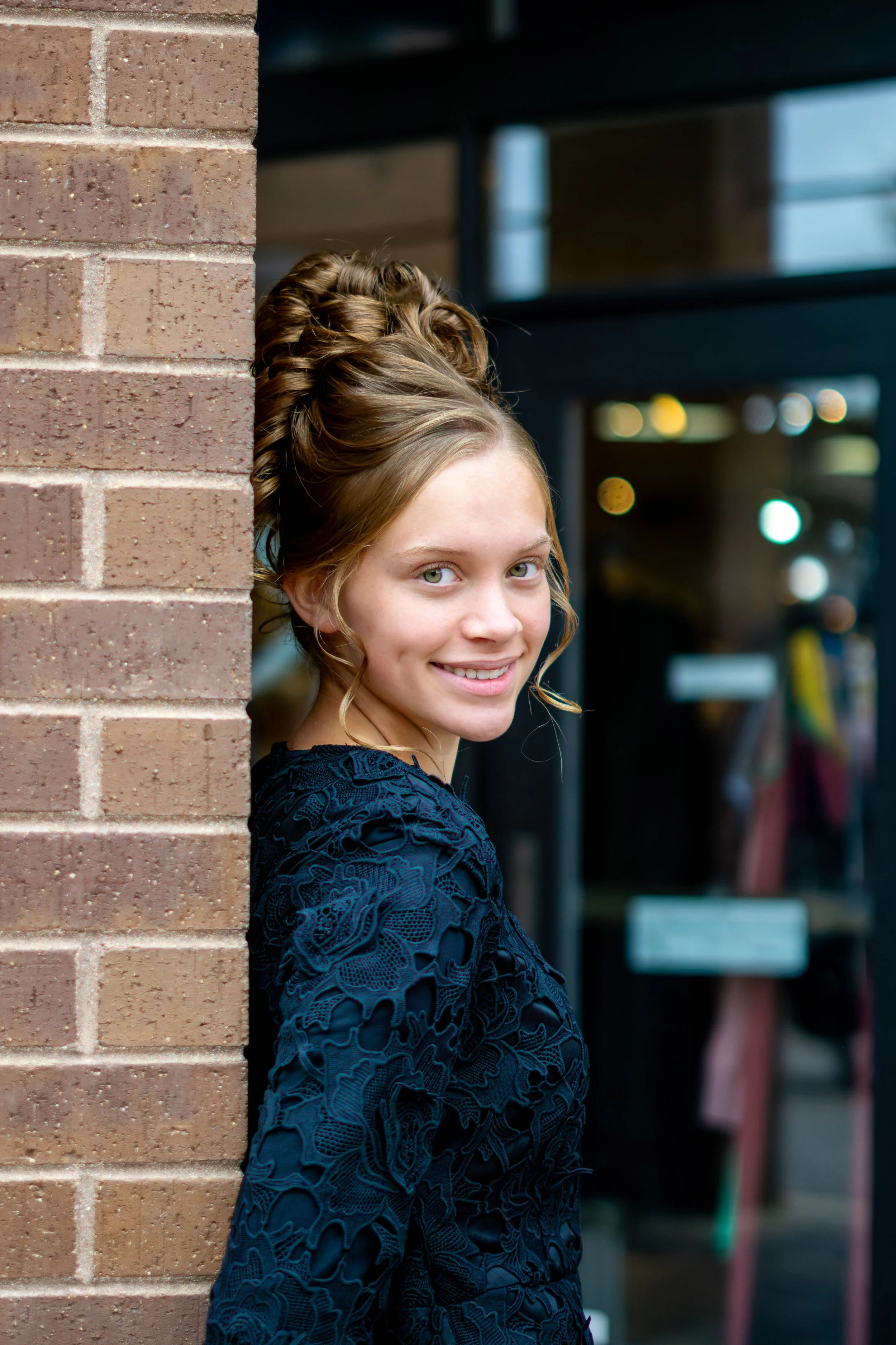 A young woman with curly blonde hair and blue eyes smiling, standing partially behind a brick wall, with shelves and windows in the background.