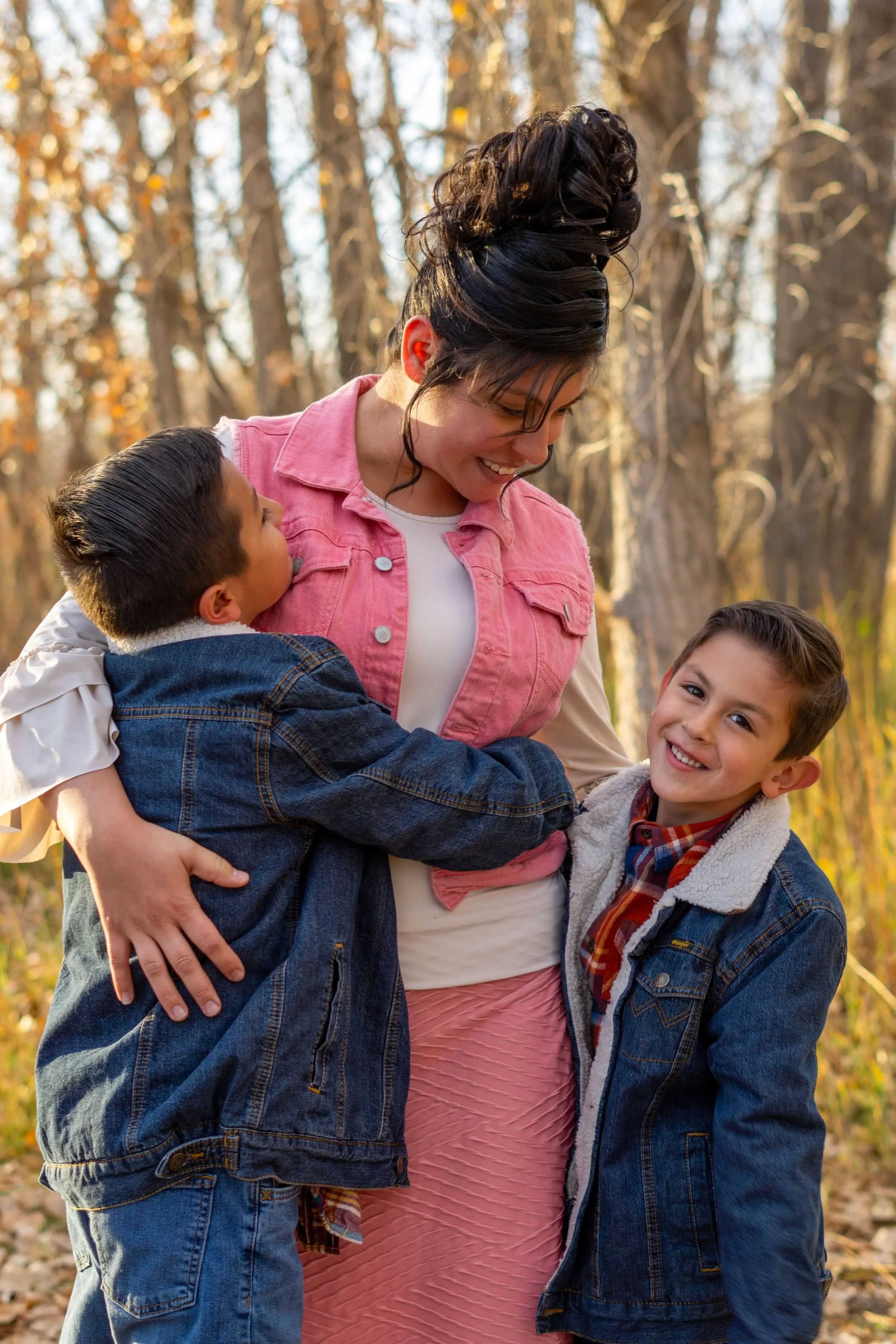 A woman with curly hair in an updo, wearing a pink jacket, hugging two young boys outdoors in a wooded area during autumn, smiling.