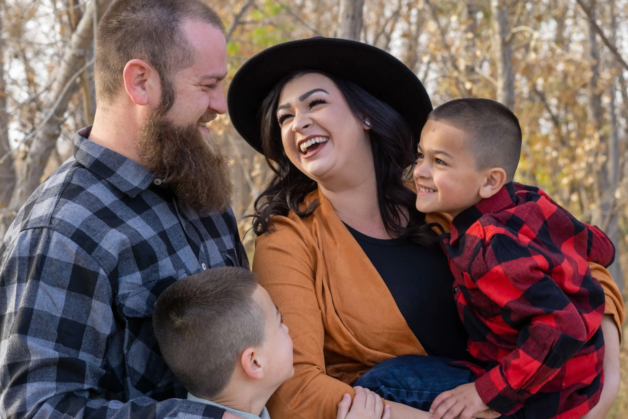 A family of five happily gathered outdoors in autumn, with trees in the background. The mother is wearing a black hat and smiling at her partner, a man with a beard wearing a checkered shirt. Their two young boys are also smiling, one in front and th