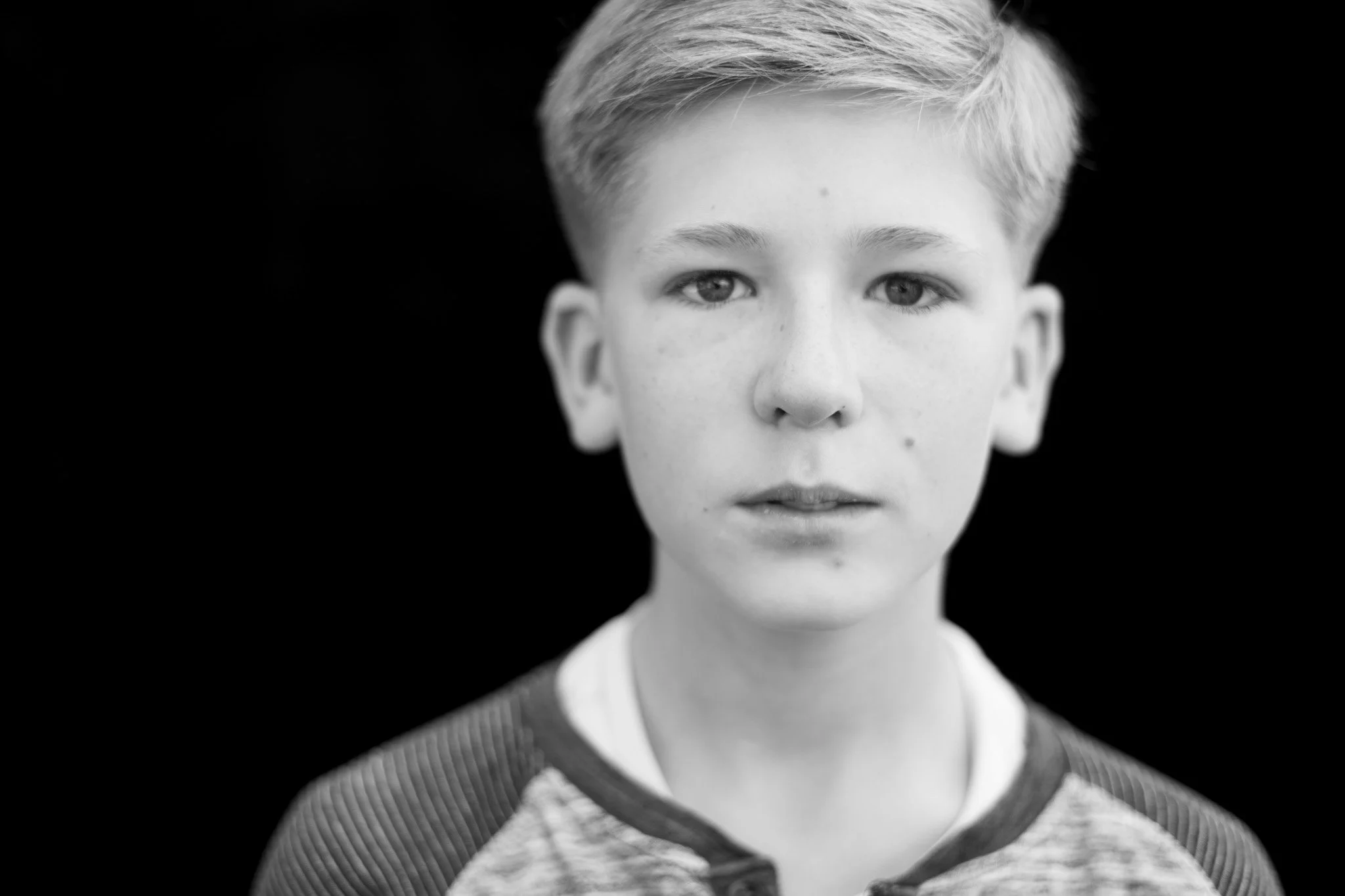 Black and white portrait of a young boy with short light-colored hair, looking directly at the camera with a neutral expression against a dark background.