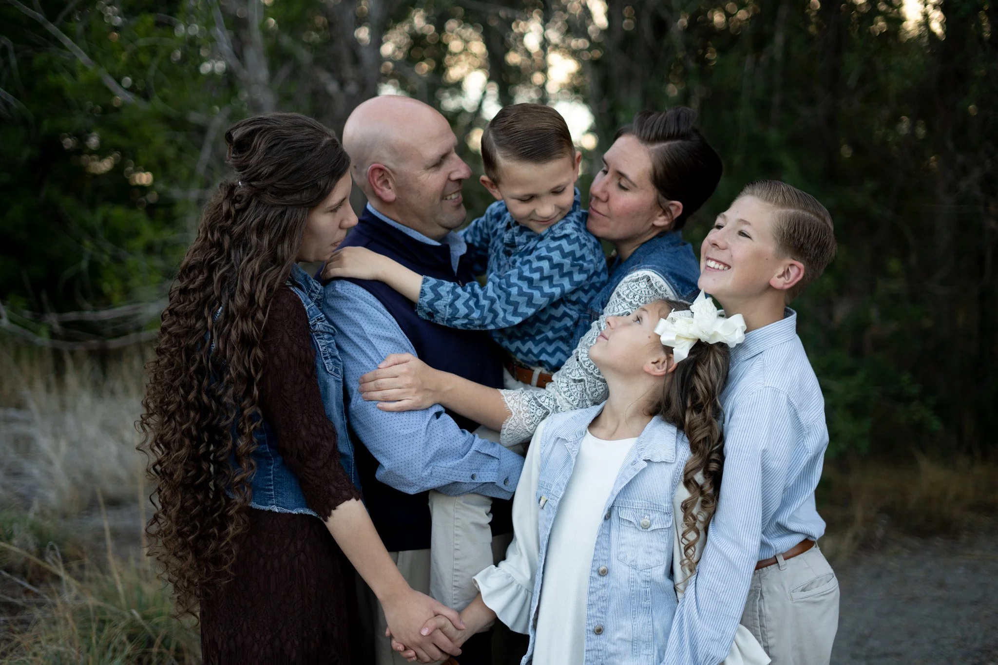 A family of six, including two adults and four children, standing outdoors in a natural setting with trees and grass, embracing and smiling at each other.
