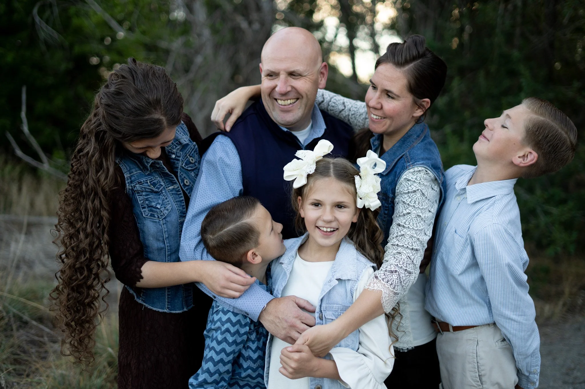 Family of seven, including three children, hugging and smiling outdoors in a park with trees in the background.