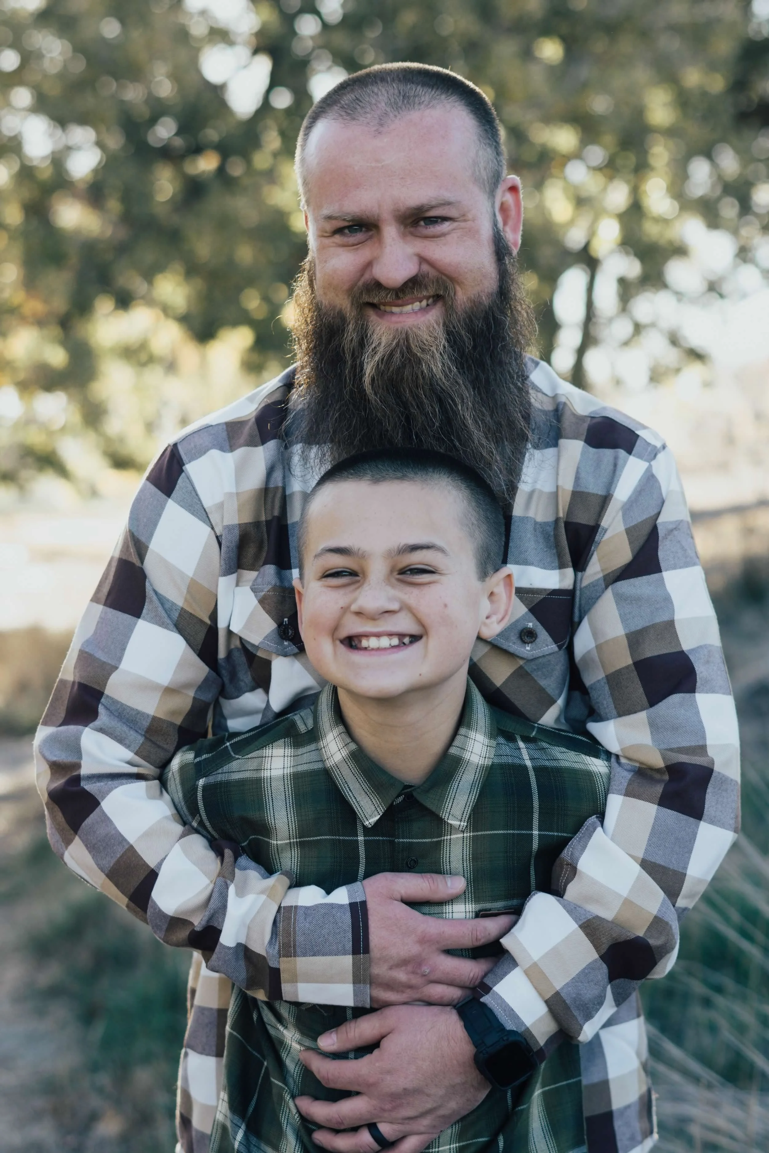 A man with a beard and a buzz cut smiling outdoors, with a boy in front of him also smiling, in a park or natural setting with trees in the background.