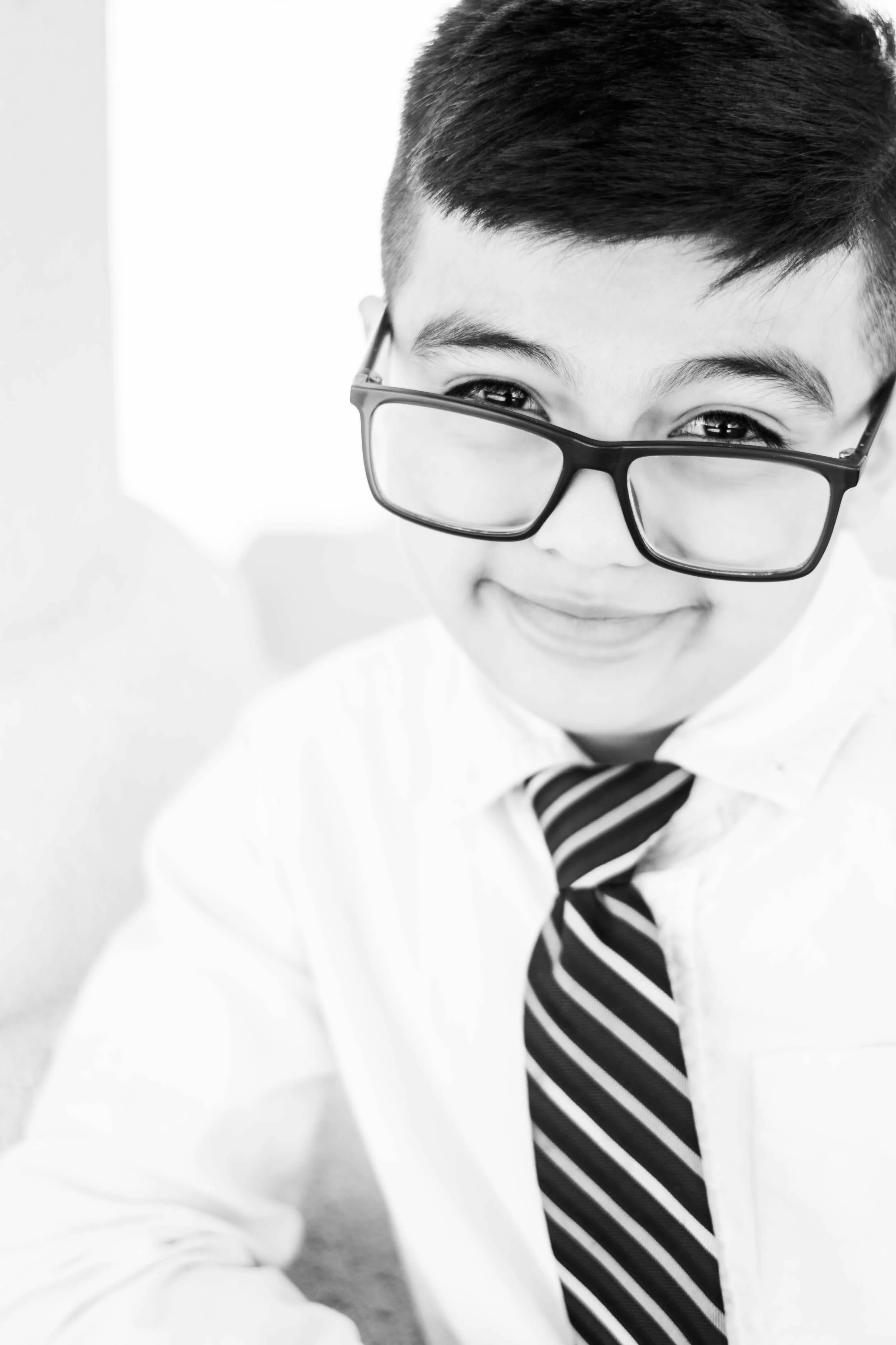 A young boy wearing glasses, a white shirt, and a striped tie, smiling and looking to the side.