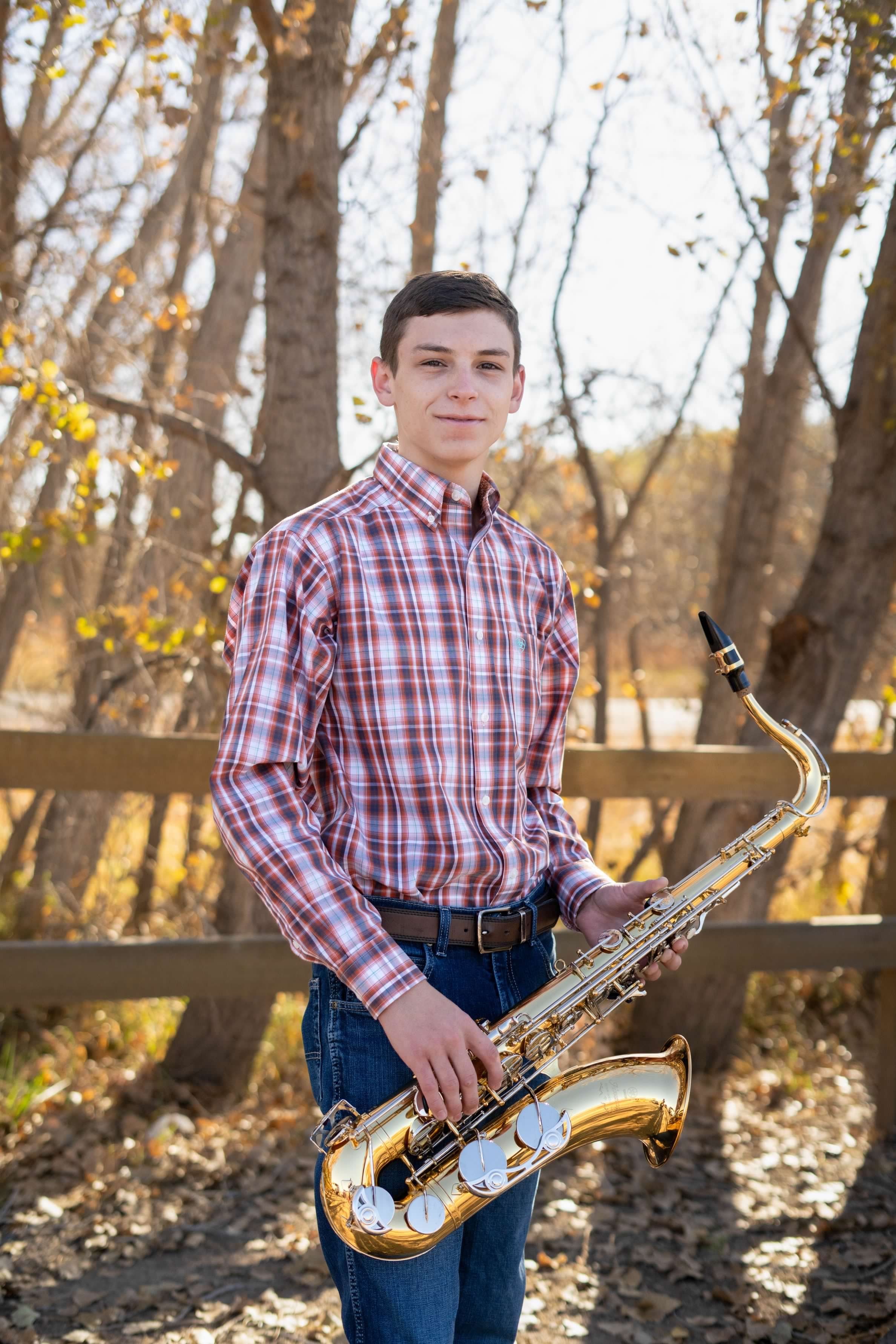 A young man with dark hair wearing a red plaid shirt and jeans, holding a gold saxophone outdoors with trees and autumn leaves in the background.