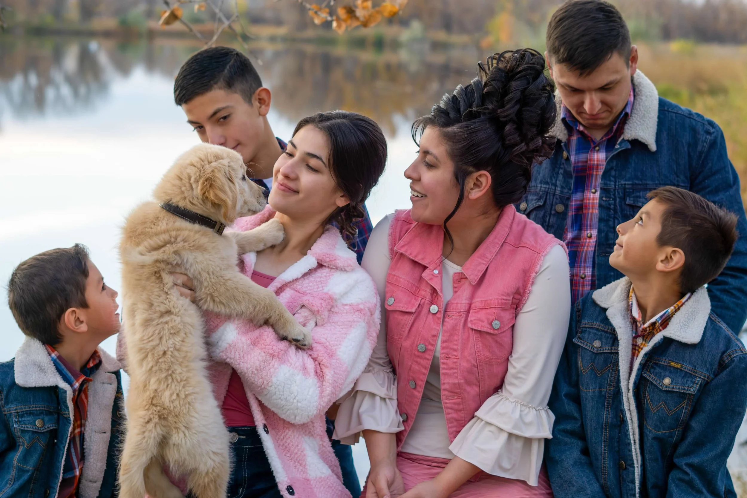 A group of children and two adults outdoors by a lake during fall, with trees and autumn leaves in the background. The children are gathered around a girl holding a light-colored puppy that she is smiling at, while the other children look on. The wom
