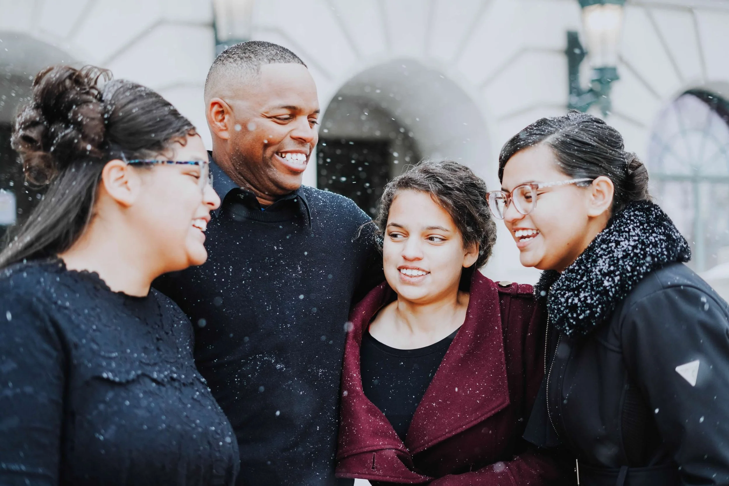 A diverse group of five people, three women and two men, smiling and laughing together outdoors in the rain, wearing dark jackets and glasses.