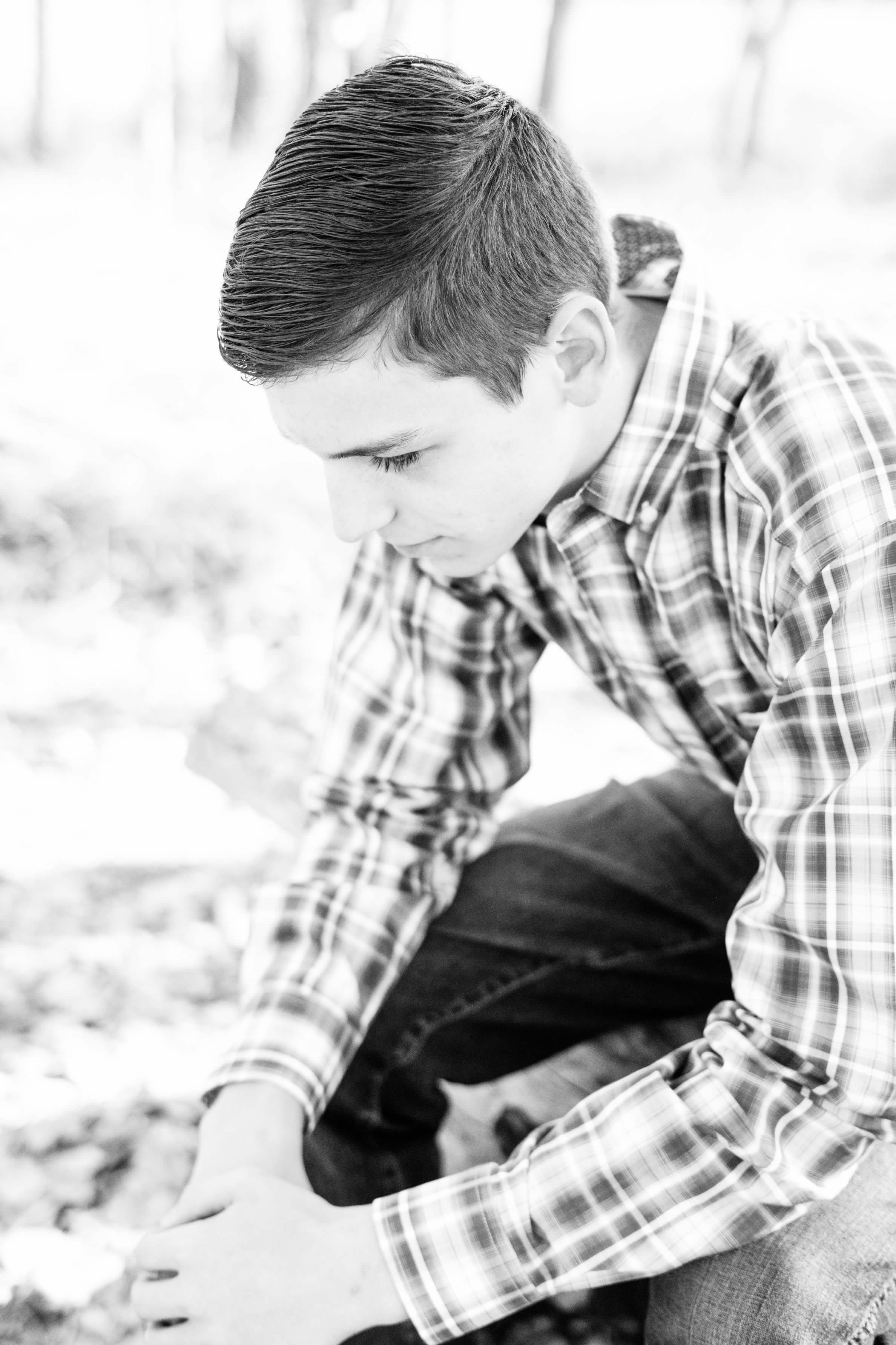 Black and white photo of a young person with short, slicked-back hair, wearing a plaid shirt, sitting outdoors with a blurred natural background.