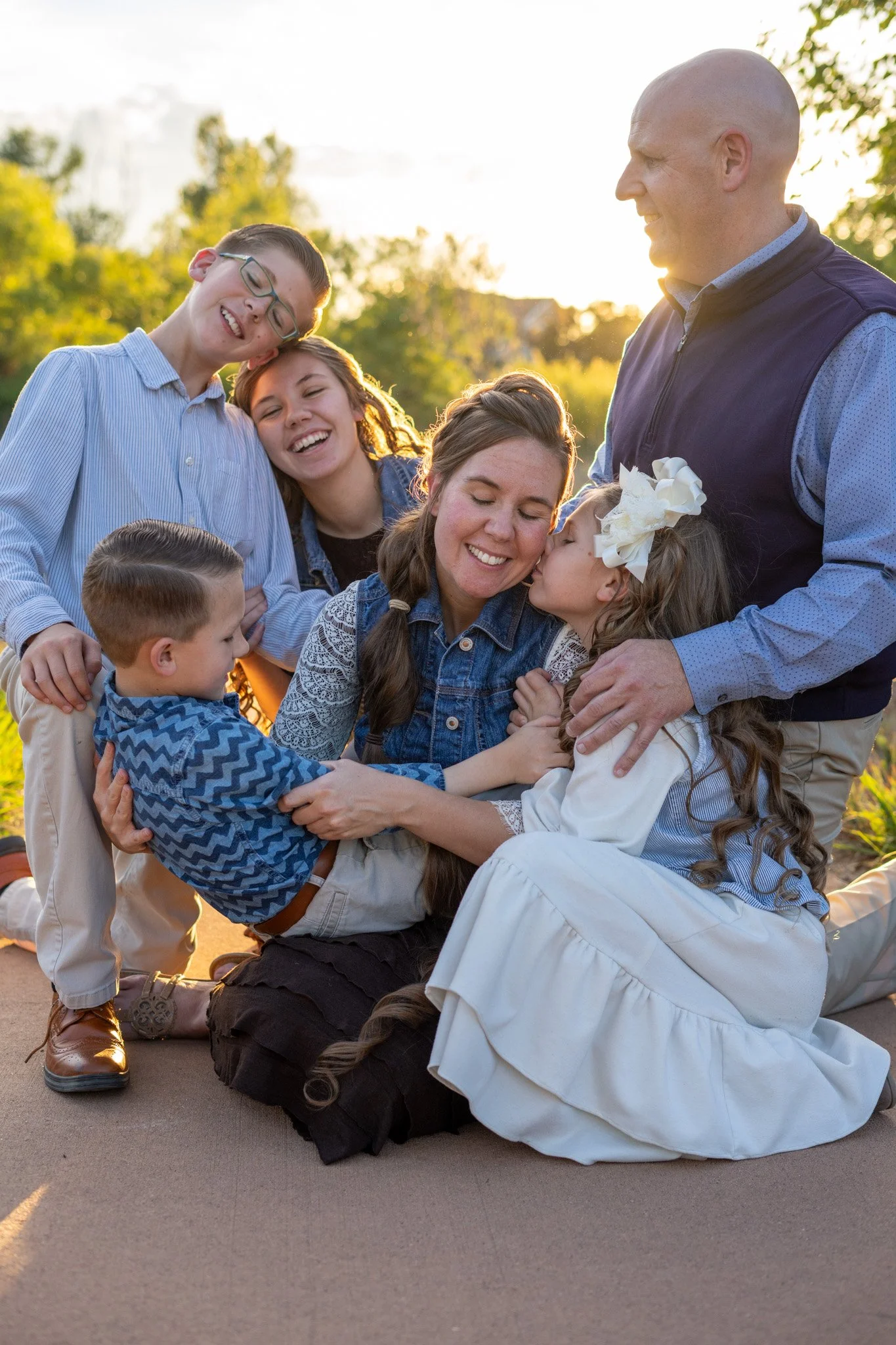 A woman and a young girl kiss while hugging on the ground, surrounded by smiling children and a man, outdoors with trees and sunlight in the background.