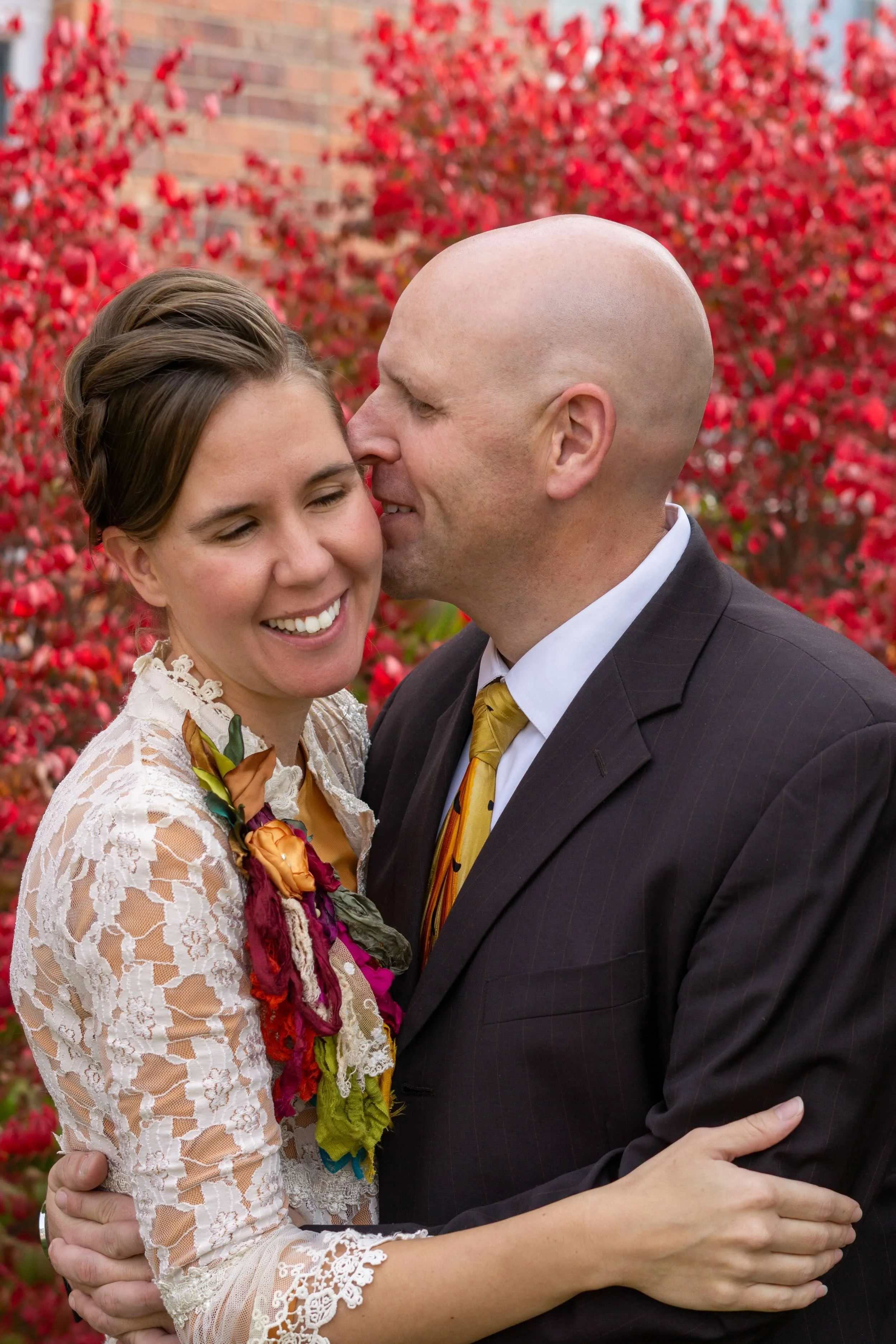 A couple hugging outdoors in front of a background of red leaves. The woman is smiling with her eyes closed, and the man is about to kiss her on the forehead. The woman is wearing a lace dress and a colorful floral accessory, while the man is dressed in a dark suit with a yellow tie.