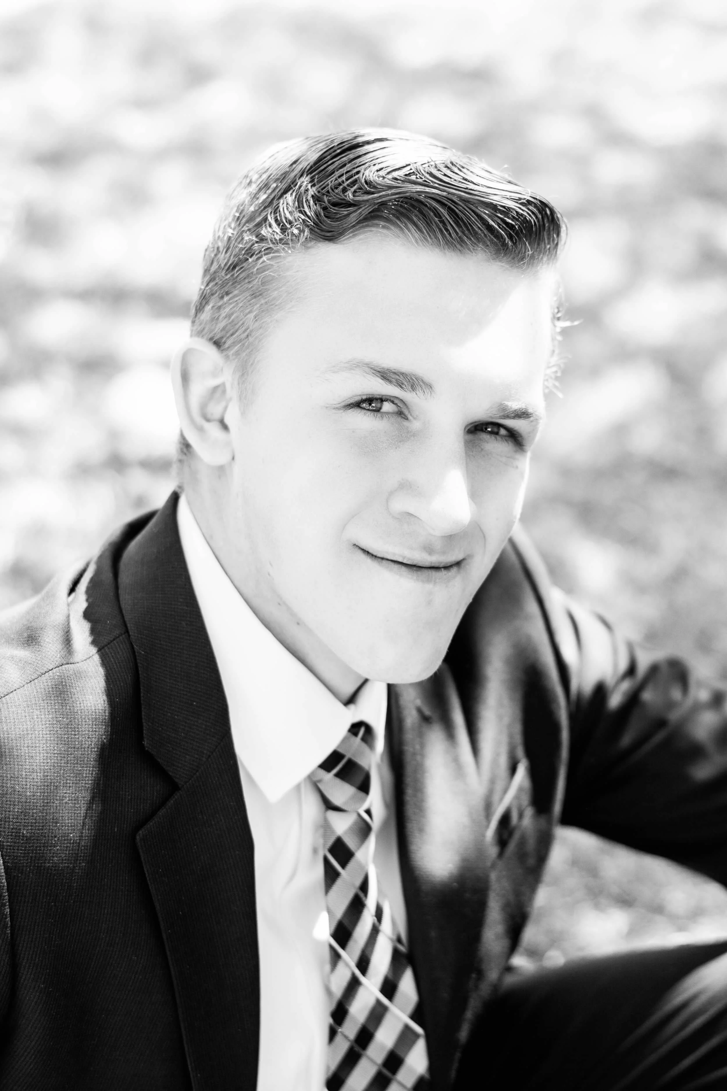 Young man with slicked back hair wearing a suit and tie, sitting outdoors.