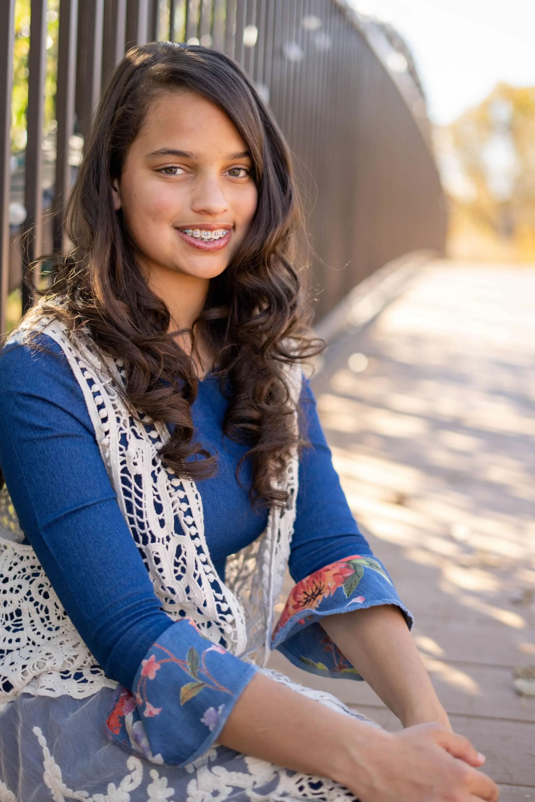 A young woman with long, wavy brown hair and braces is sitting outdoors on a wooden bench next to a metal fence during daytime. She is wearing a blue long-sleeve dress with floral patterns on the sleeves and a white lace vest. The background shows blurred trees with autumn-colored leaves and bright sunlight.