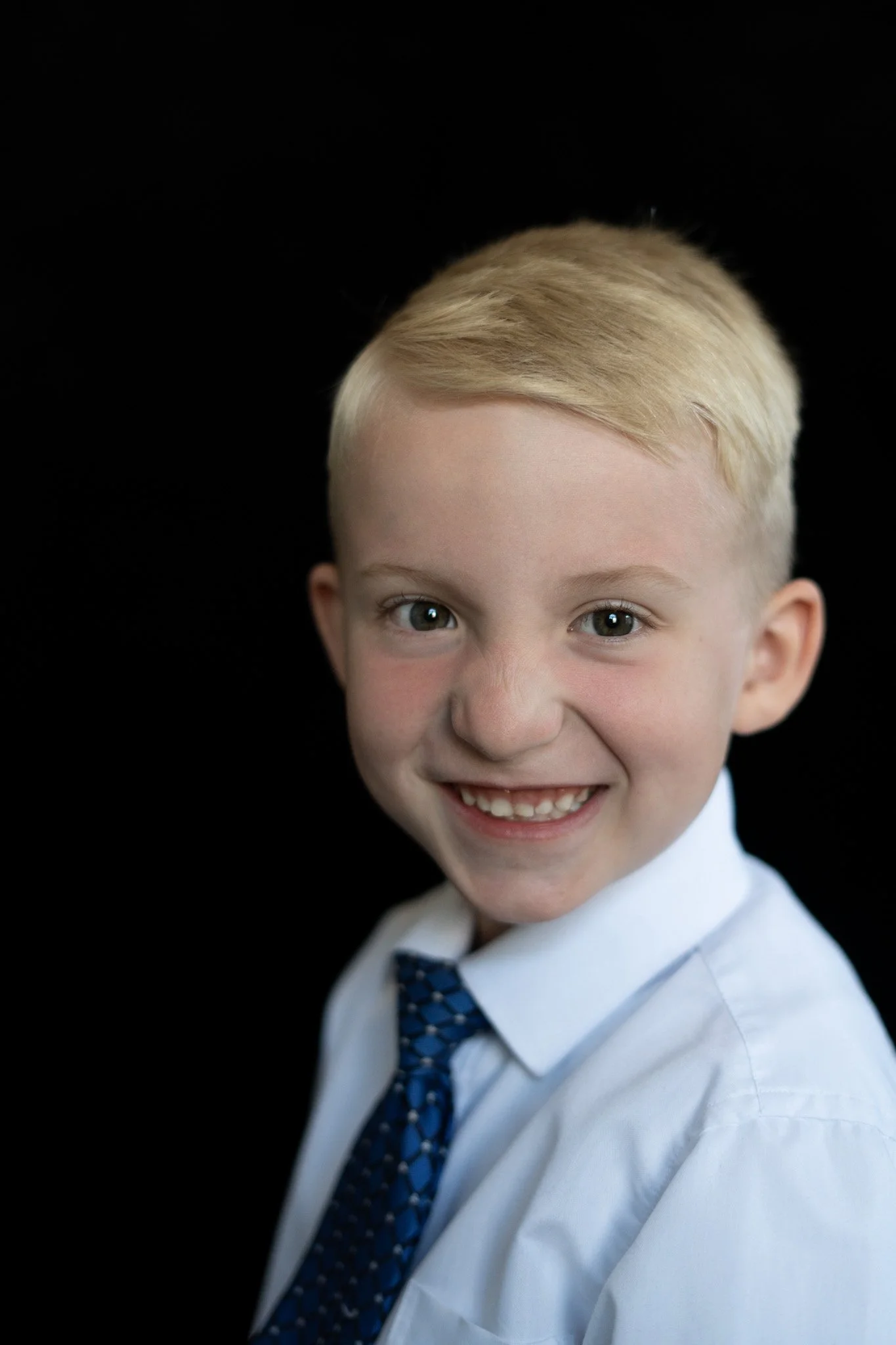 A young boy with blonde hair, blue eyes, and a big smile, dressed in a white shirt and a blue patterned tie, looking directly at the camera with a black background.