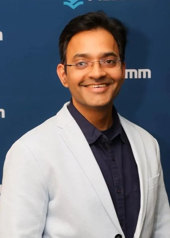 Sushant Dayal wearing a white blazer over a dark shirt, standing in front of a blue background with logos.