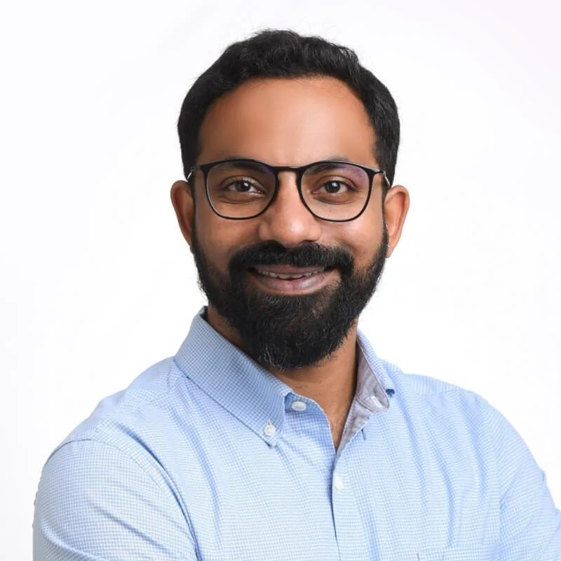 Aditya smiling man with glasses and a beard wearing a light blue collared shirt, posing against a plain white background.
