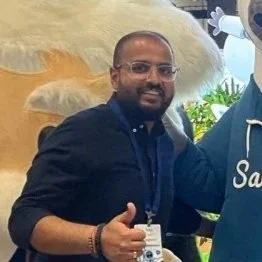 Pulkit with glasses, a beard, and a dark shirt smiling and giving a thumbs-up at an outdoor event, with a decorative lion statue in the background.