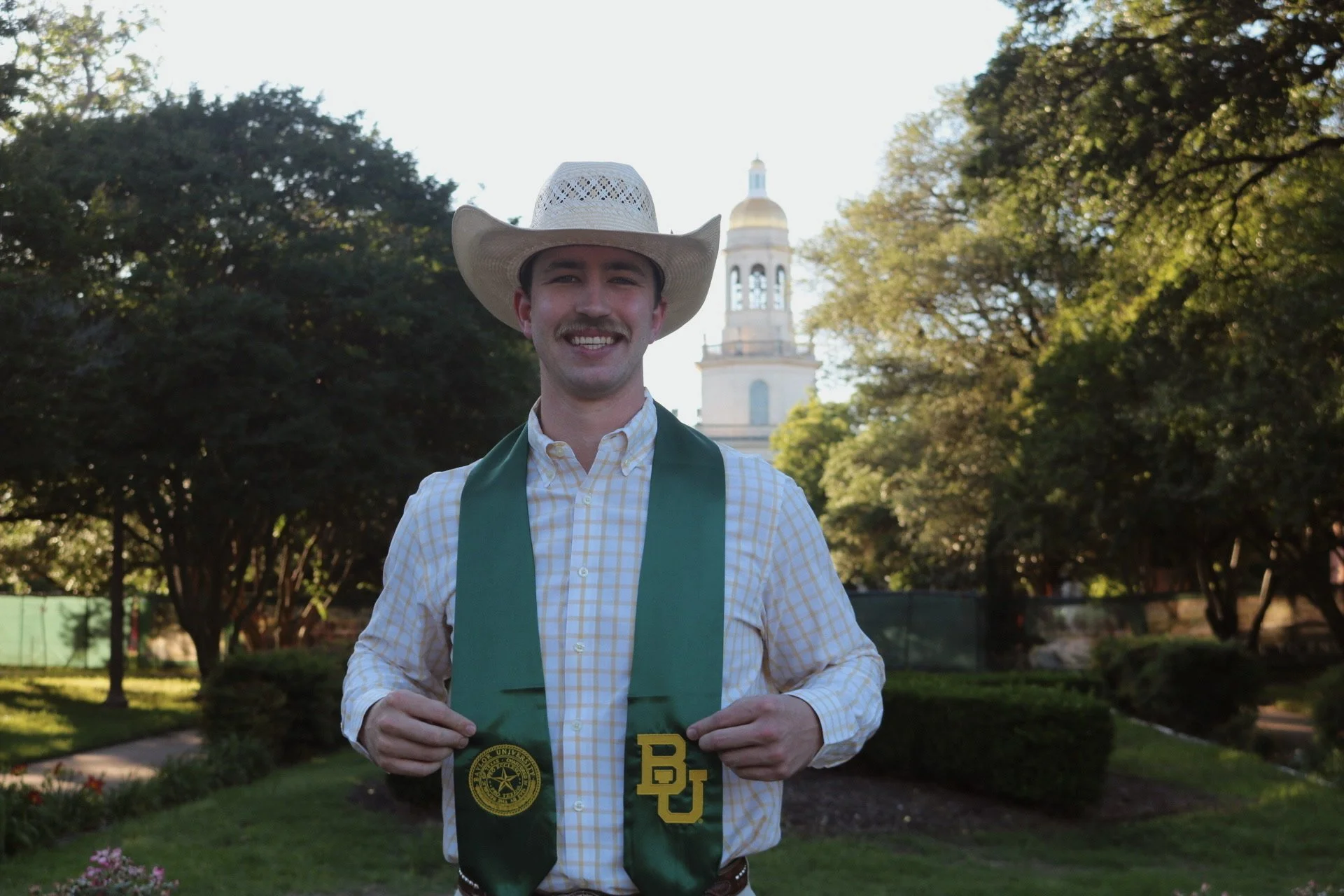 A man in a cowboy hat and checked shirt standing outdoors, holding a green stole with university logos in front of a historic building and trees.