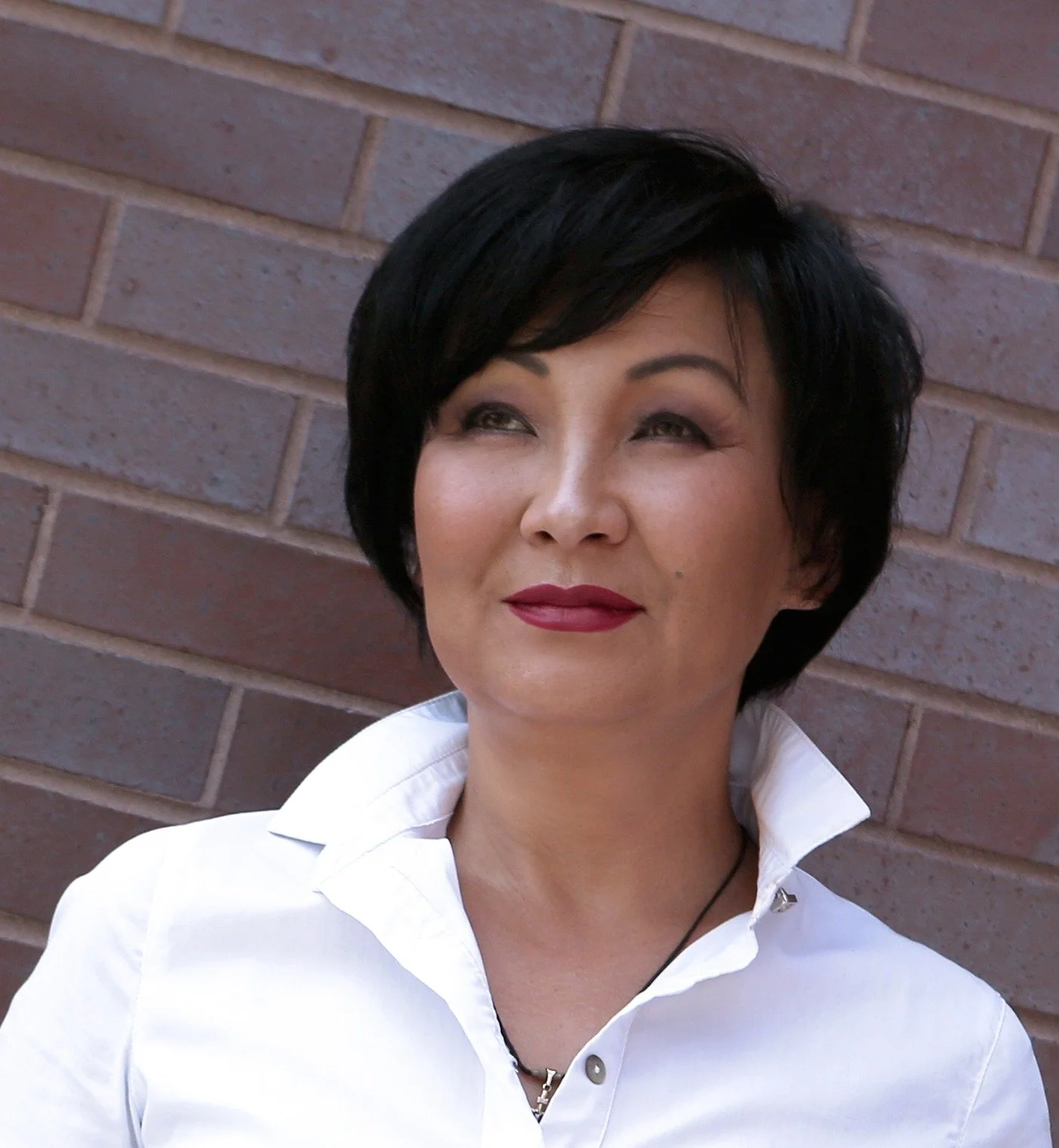A woman with short black hair, wearing a white collared shirt, standing against a brick wall.