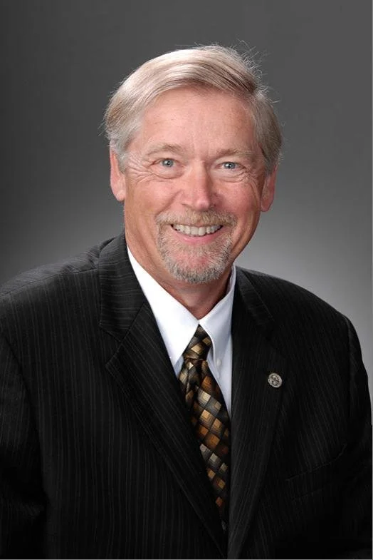 Professional portrait of Gene Krebs man with gray hair and a beard, wearing a dark pinstripe suit, white shirt, and patterned tie, against a plain gray background.